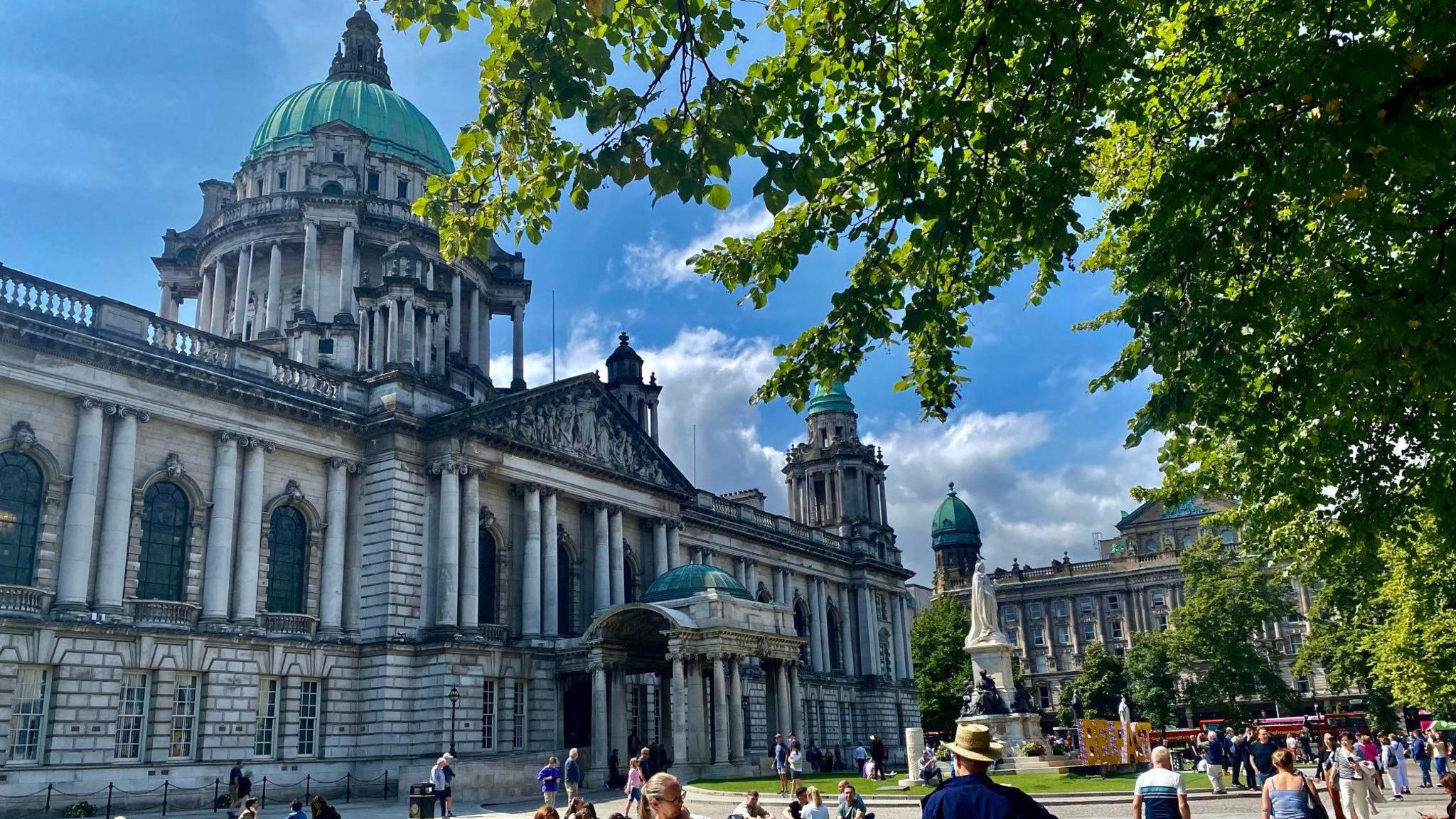 Belfast city hall on a sunny day. The building is grey and there are lots of people walking around the gardens of it.