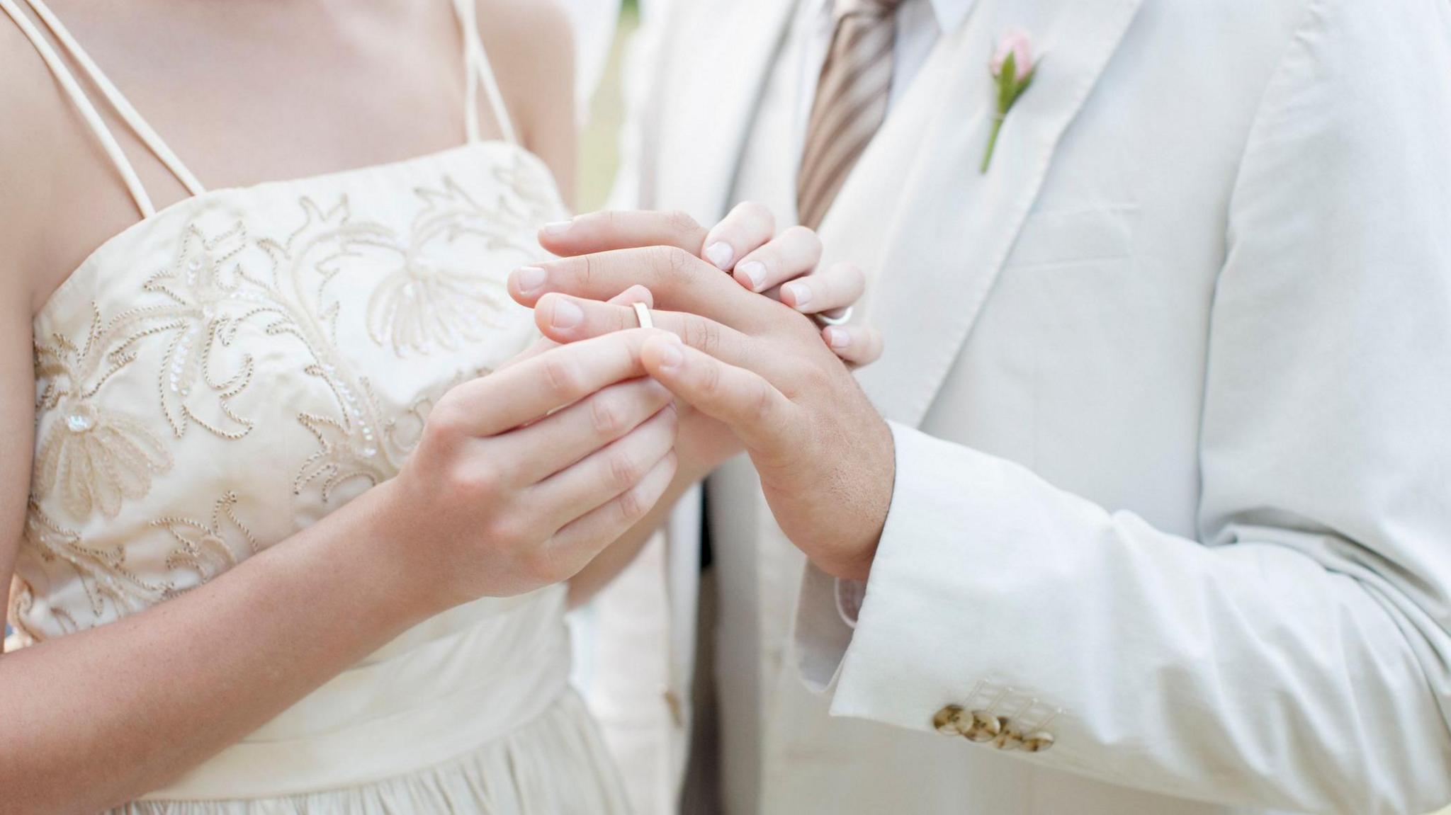 A woman in a white dress puts a ring on the finger of a man wearing a white suit