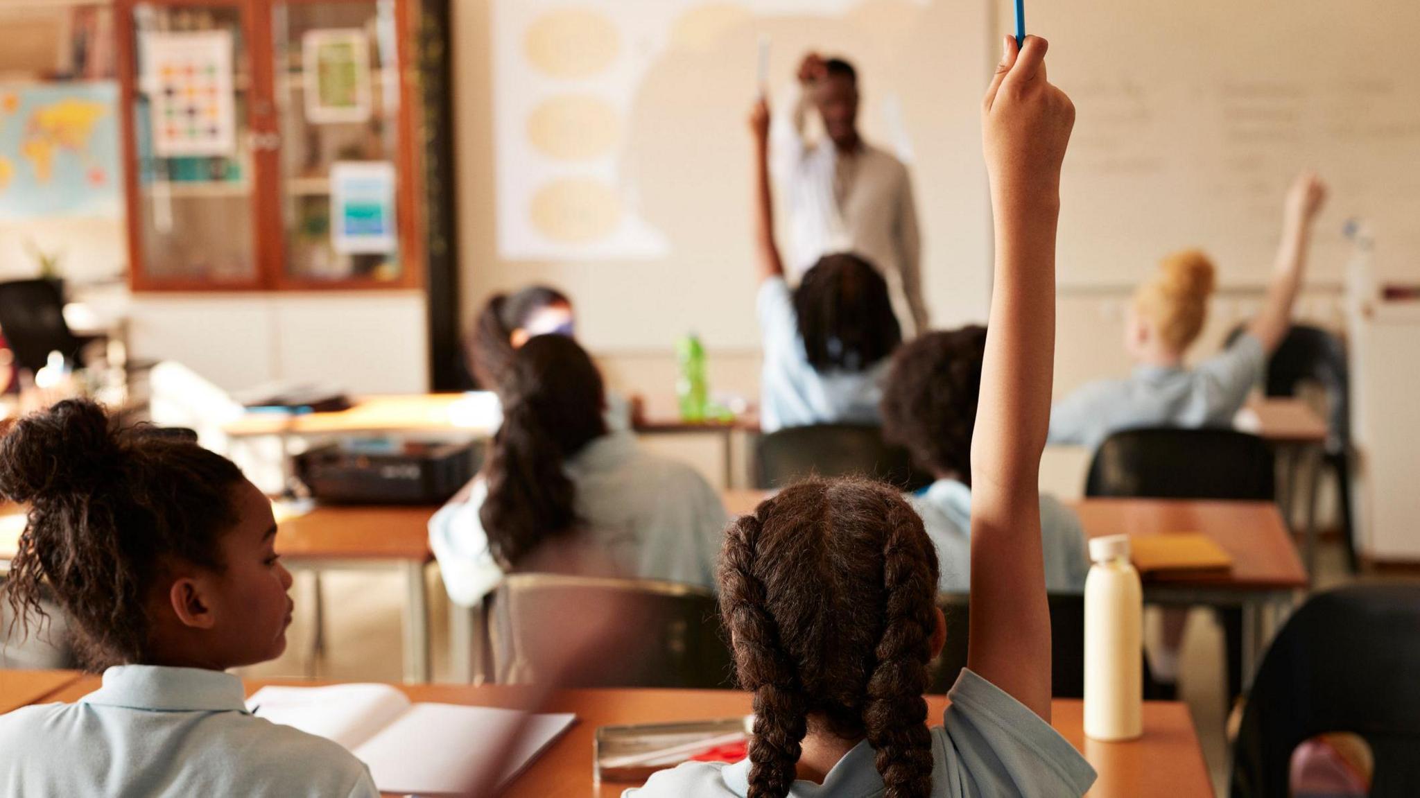 Children in classroom, some with raised arms. 