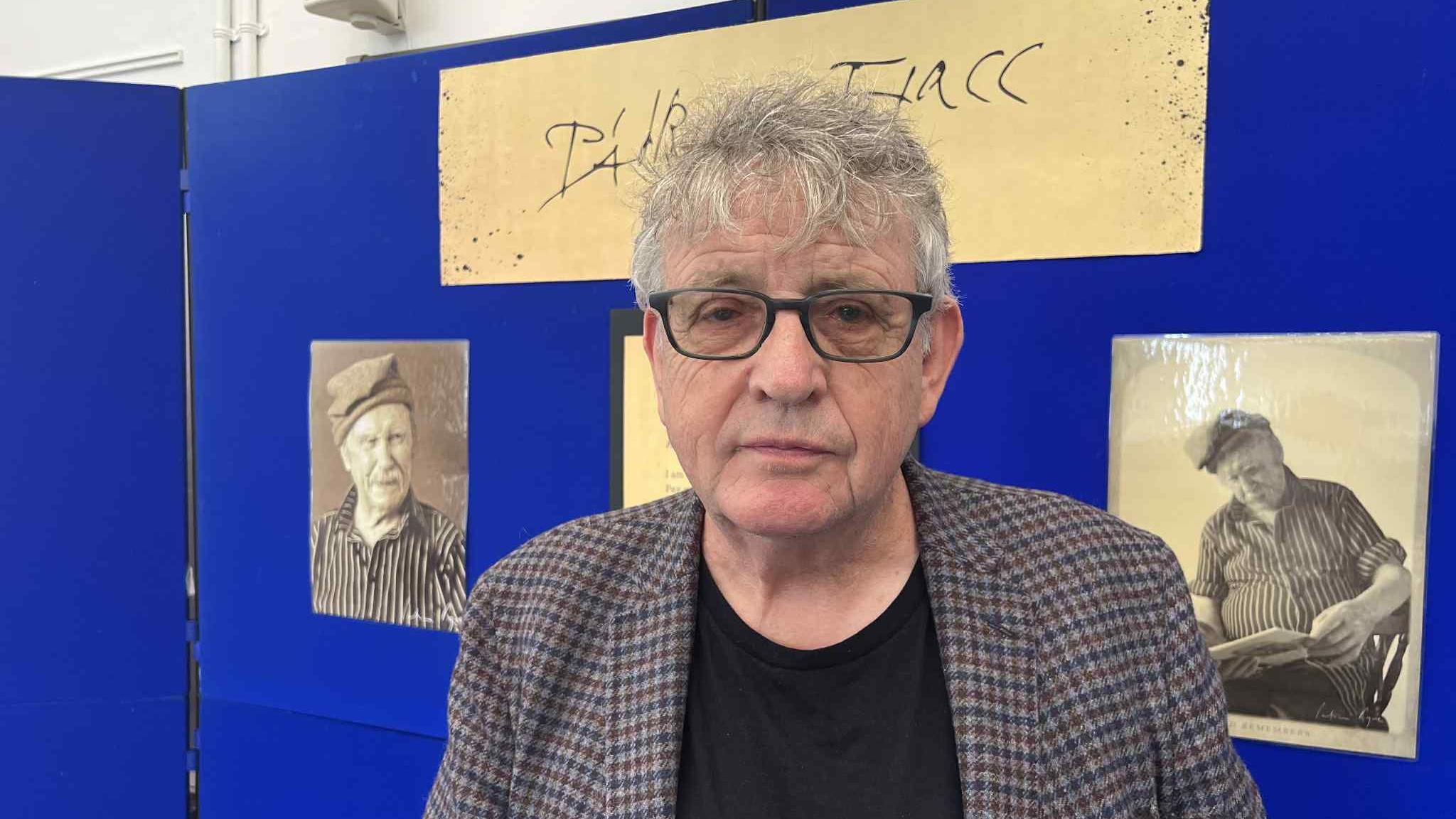 Paul Muldoon has medium-length grey hair and is wearing dark-rimmed glasses and a grey, blue and brown checked suit jacket over a black T-shirt. Behind him are black and white photos of Padriac Fiacc on a blue noticeboard.