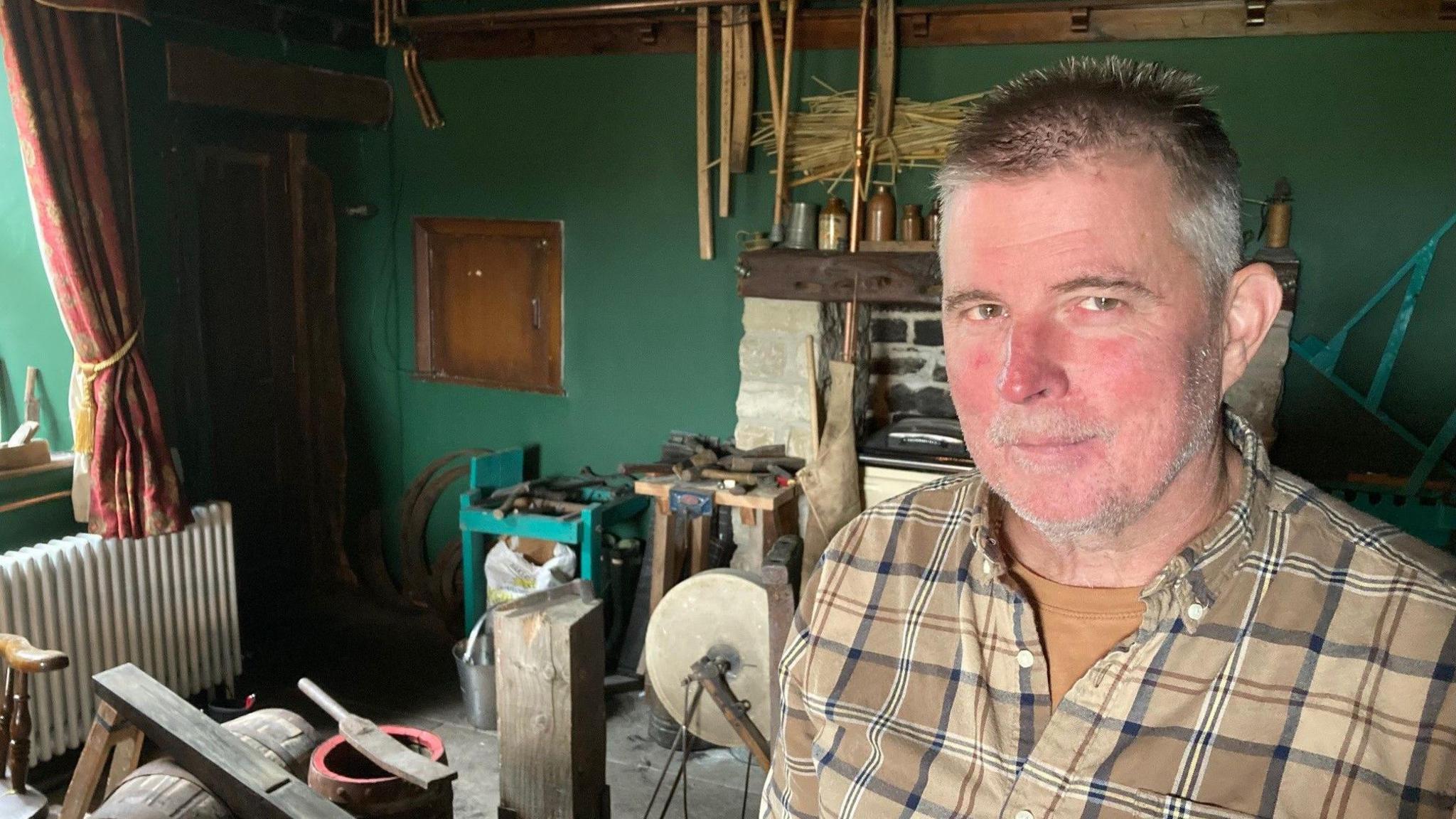 A white-skinned man with ruddy coloured cheeks wearing a checked shirt with wooden barrel-making equipment in the background including a partially made barrel and a round sharpening stone on a metal axle.  