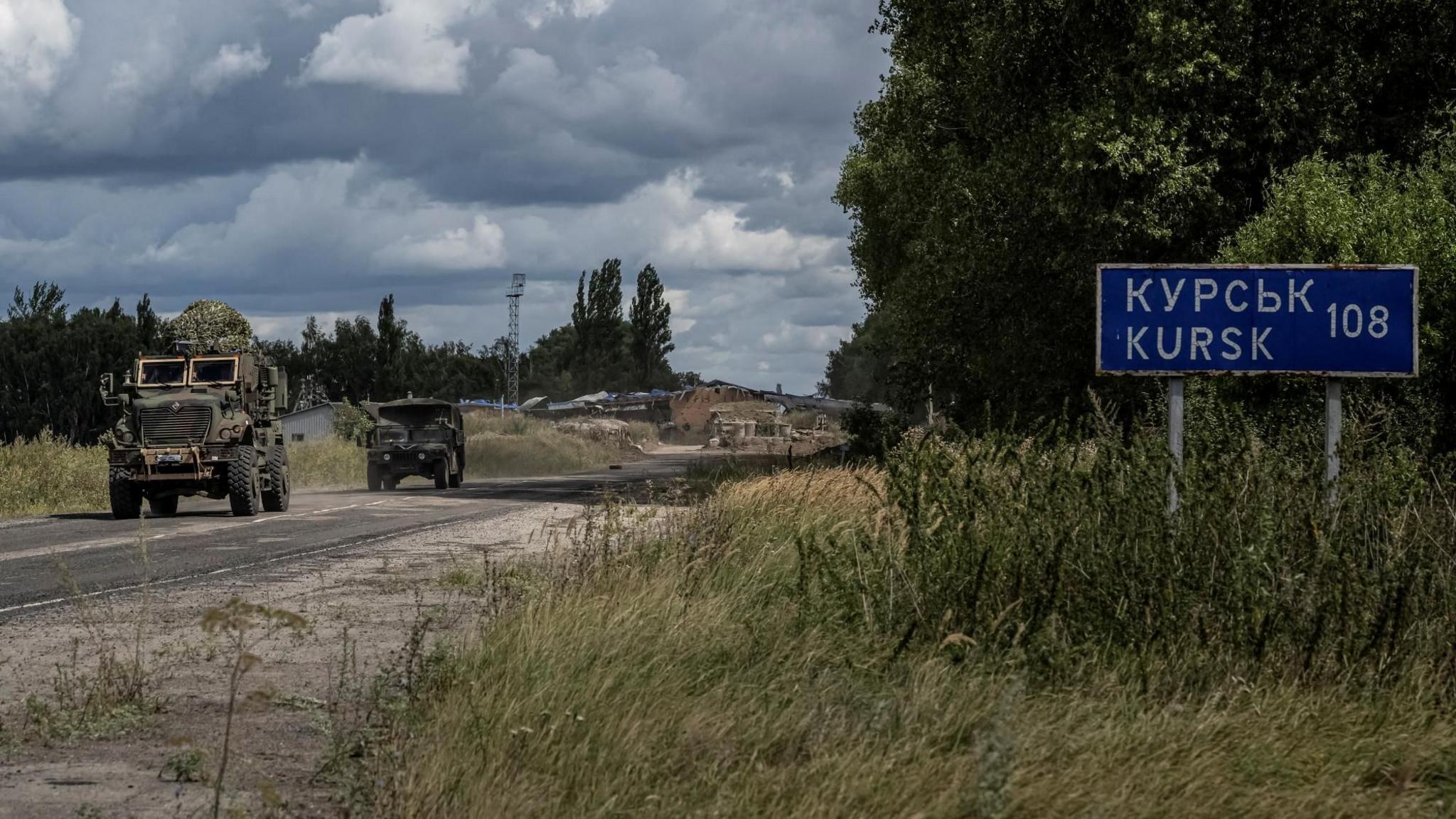 Two military vehicles driving on a road past a sign reading 'Kursk'