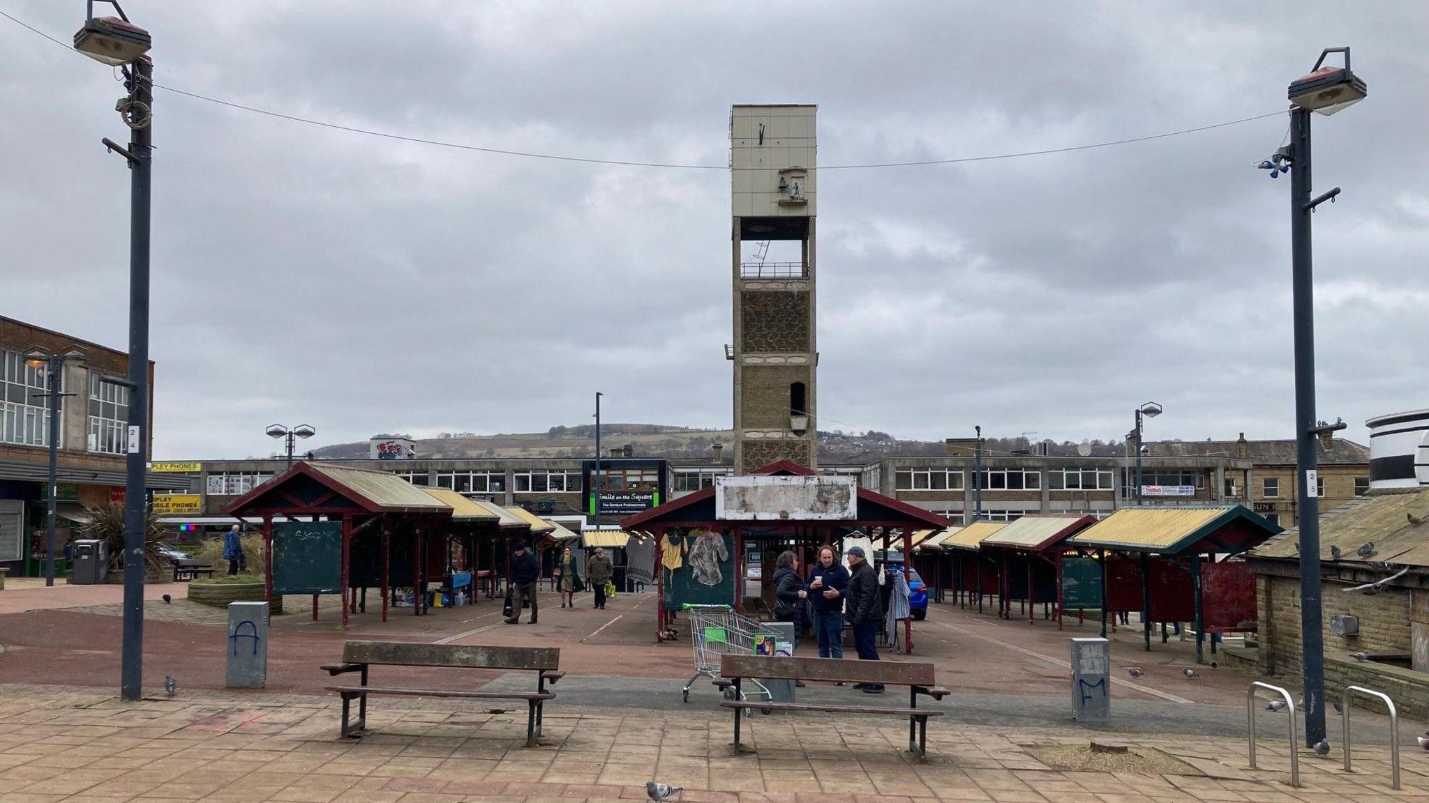 Three rows of fixed metal market stalls with a concrete clock tower in the background.