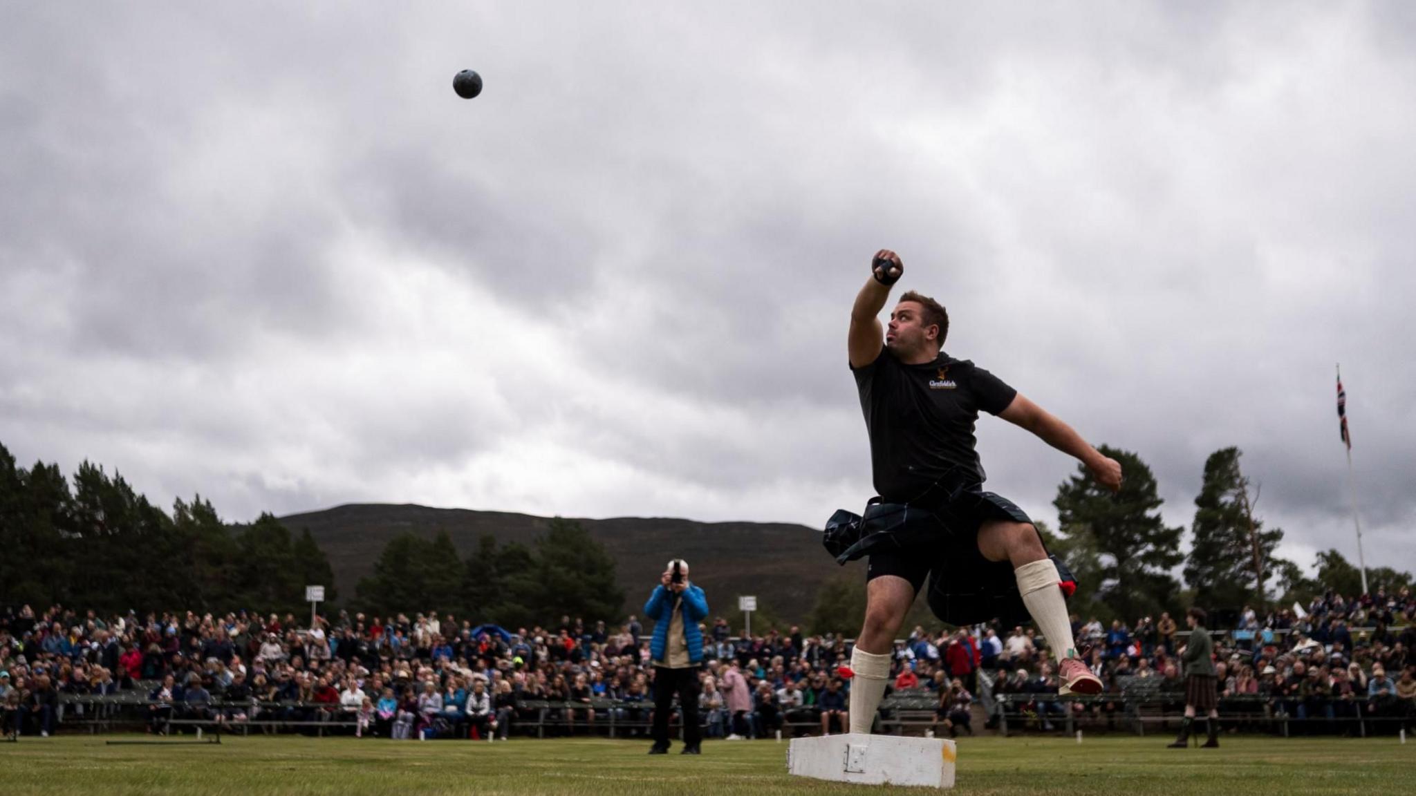A man taking part in a shot put event. He is shown in mid-action, having just released the shot put ball, which is visible in the air. The setting is outdoors on a grassy field, with a large crowd of spectators gathered in the background. Beyond the crowd, there are trees and hills, and the sky is overcast, filled with clouds.