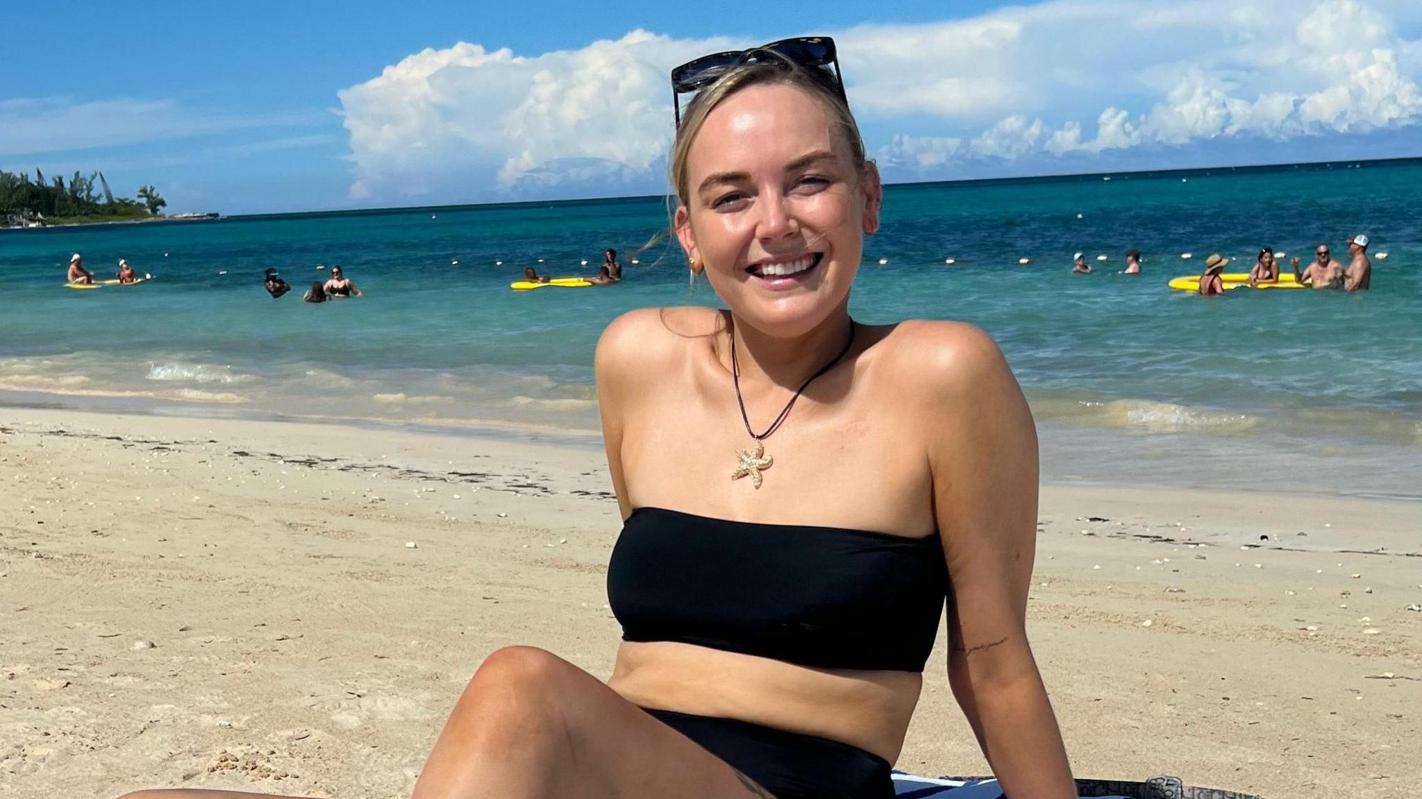 Georgie in a black bikini on a beach, smiling at the camera with the sea behind her