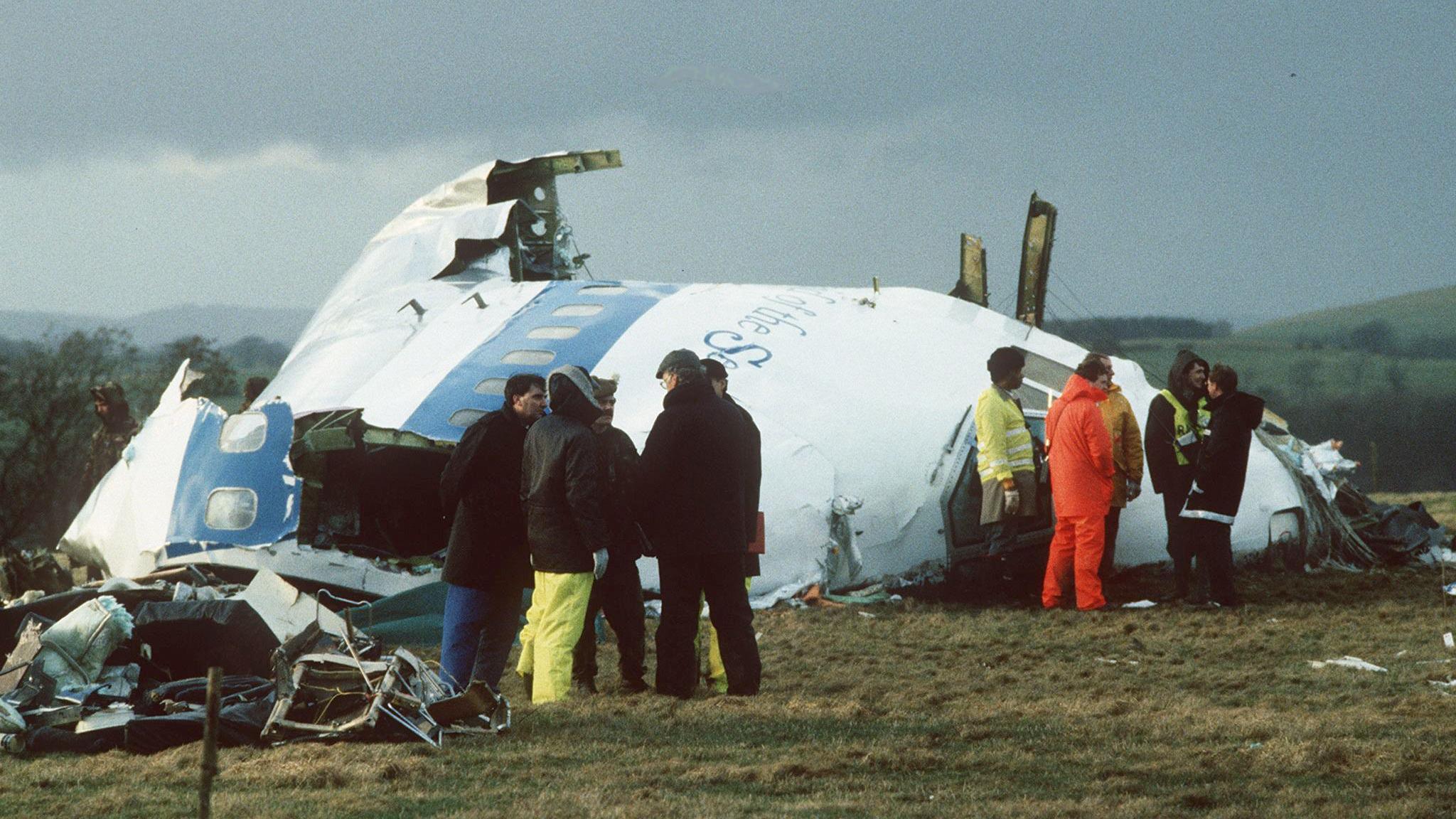 Emergency workers survey some of the wreckage of Pan Am Flight 103. There are several people standing around some of the wreckage of the plane.