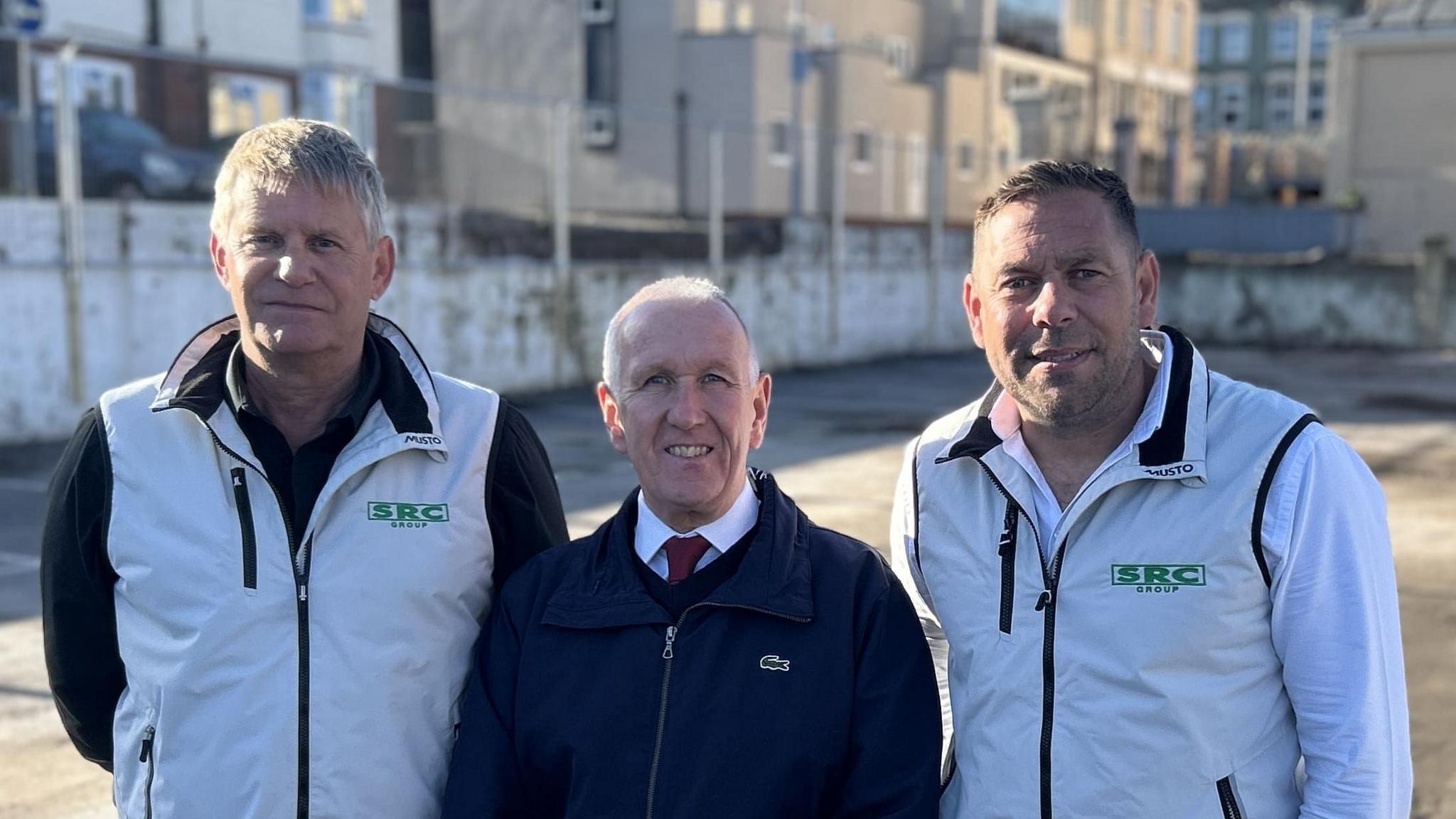Ivan Henderson is flanked by two men who worked on the car park demolition. They are wearing white jackets and Henderson is wearing a red jacket with a white shirt and red tie underneath.
