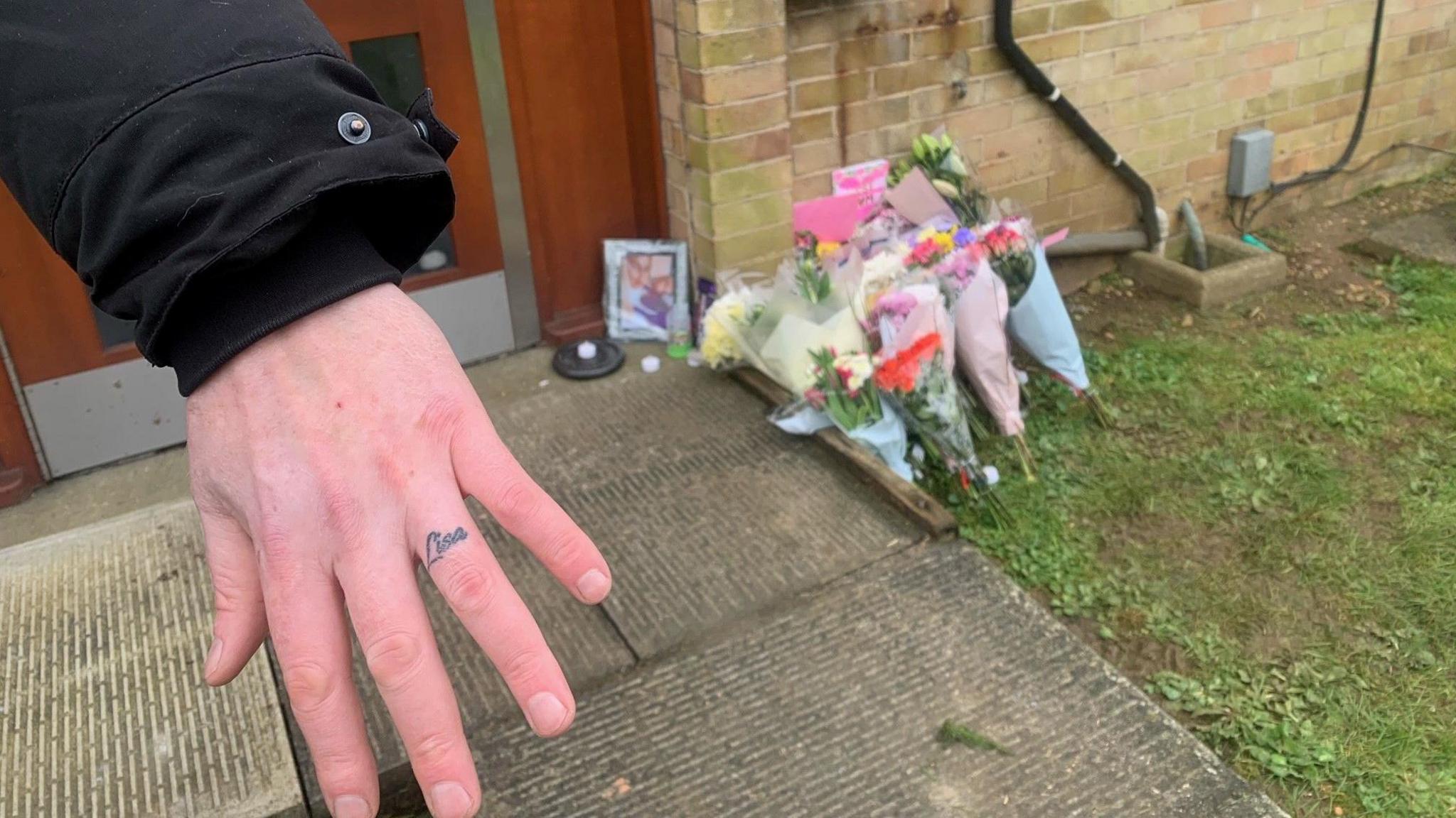 Ben wth his Lisa tattoo on his hand in front of flowers placed outside the victim's house 