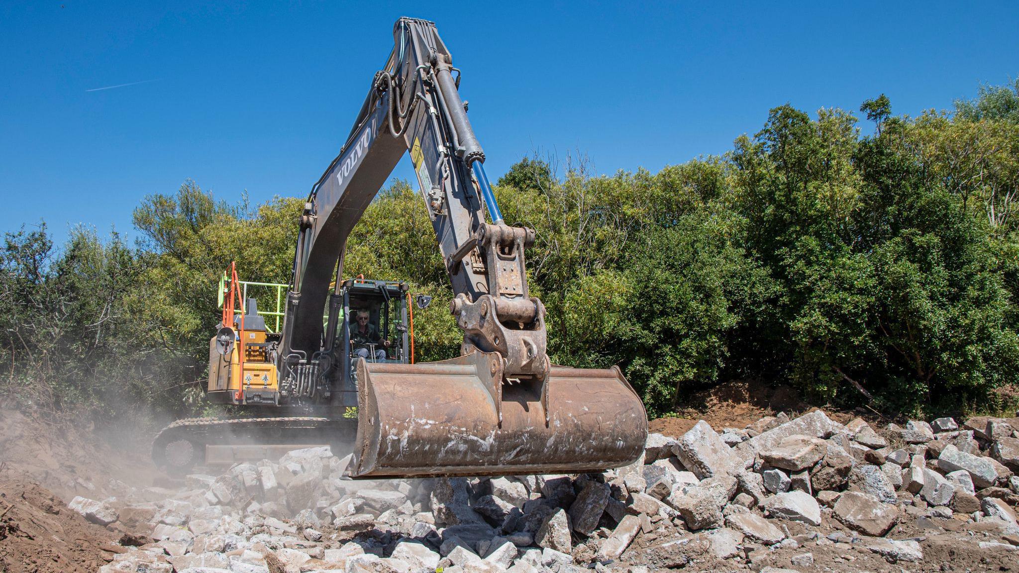 A digger lifting and moving lots of heavy rocks and debris with its extended arm. There are lots of trees and greenery in the background.