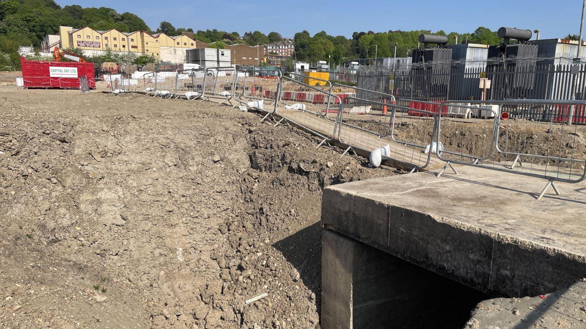Development with buildings in the distance. Barriers frame the site which is made up of bare rock and rubble.