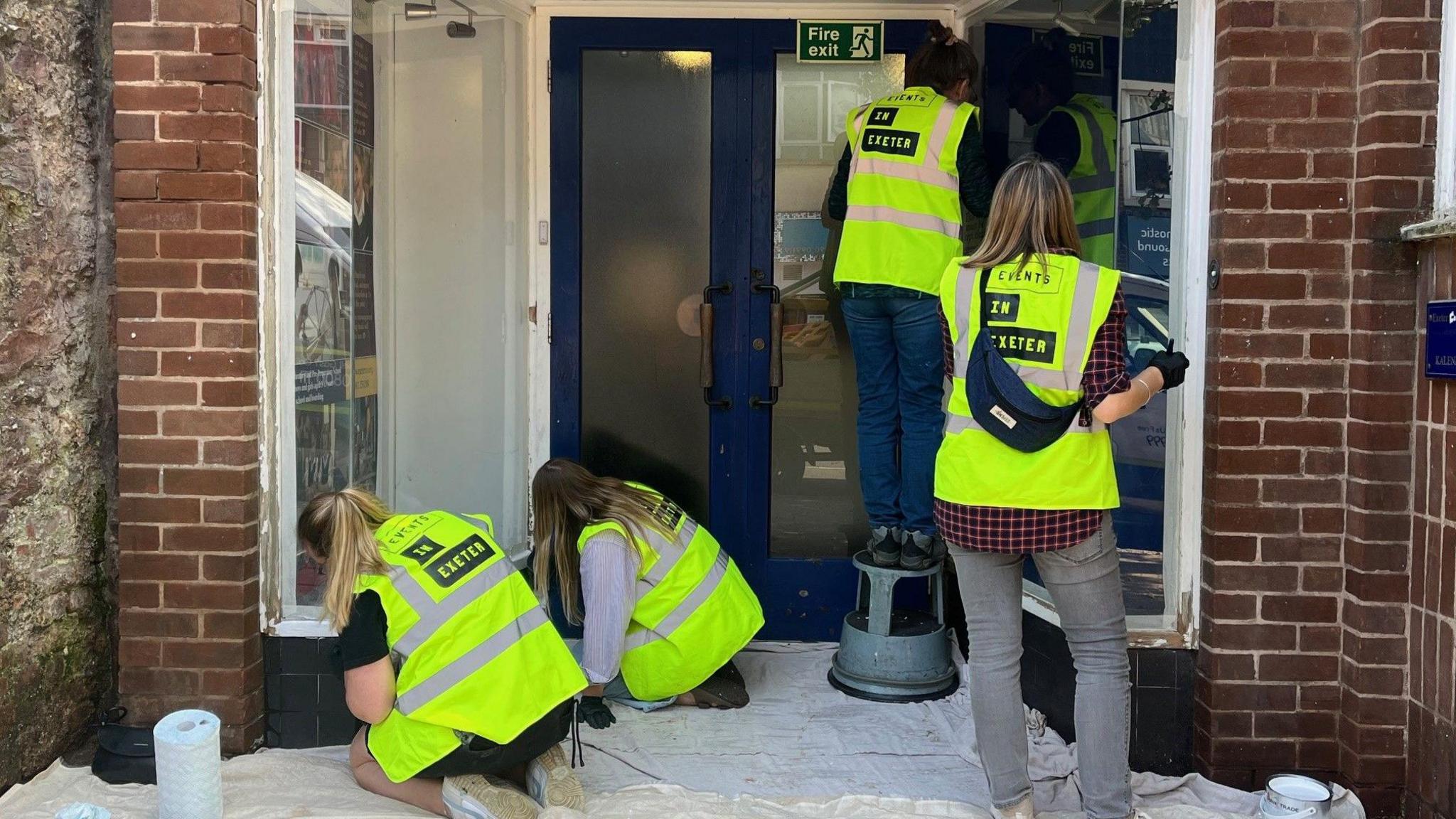 Image shows a group of four volunteers painting a doorway in South Street, Exeter. The group is made up of four women with blonde or brown hair.