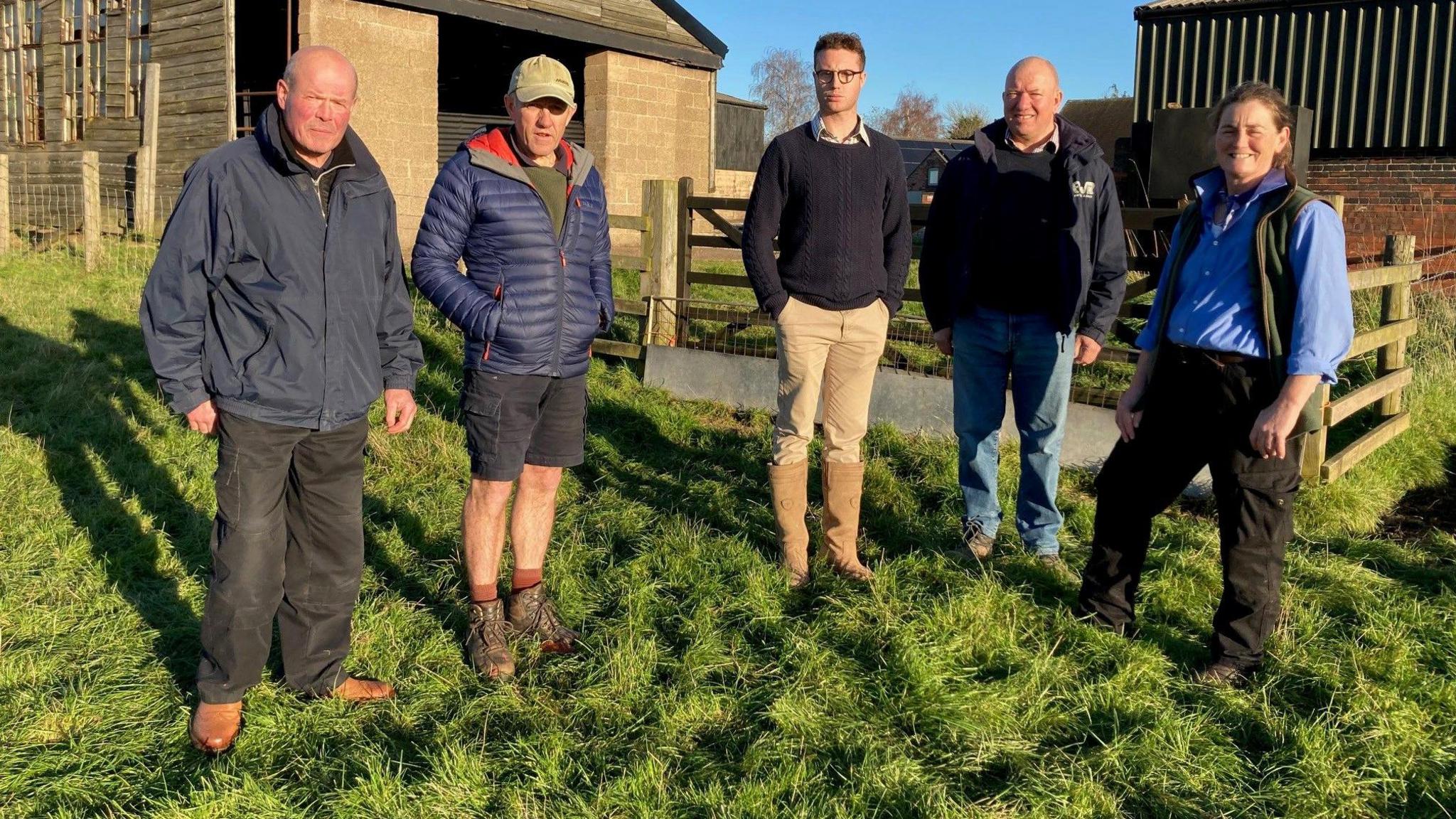 Five farmers from Staffordshire and Derbyshire standing in front of barns. On the left is a balding man with grey hair who wears a dark blue jacket and dark trousers with brown shoes. Next to him is a man with dark shorts, green boots and a puffy blue jacket over a green top with a green baseball hat on his head. In the middle is a man with dark hair, glasses, a dark jumper, light brown trousers and brown boots. Next to him is a bald man with a dark blue jacket over a dark jumper with blue trousers. Lastly, on the right is a woman with dark brown hair, a green waistcoat over a light blue shirt and dark trousers. They stand on green grass by several farm buildings made of concrete blocks and corrugated metal with a wooden fence between them and the buildings.