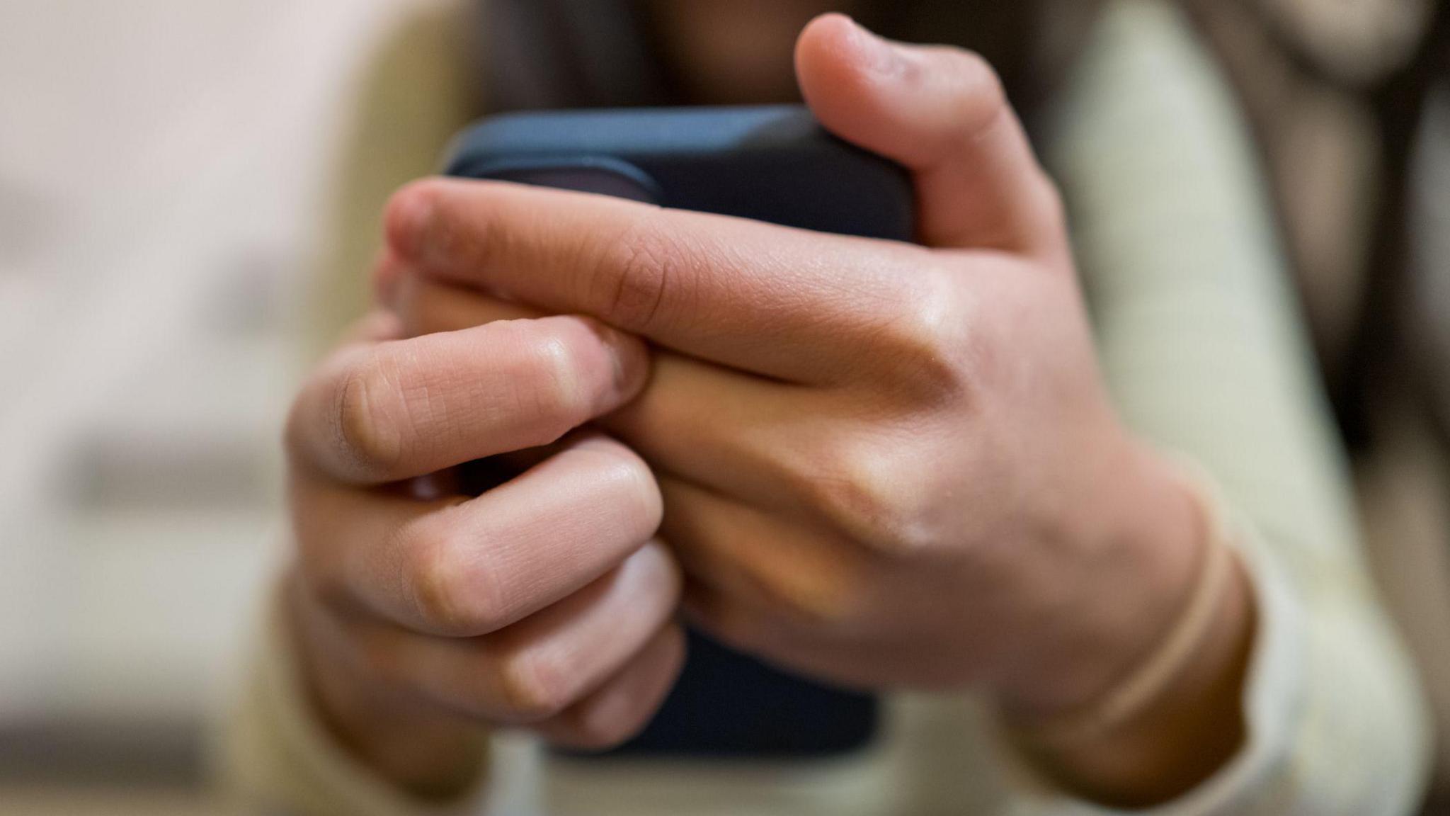 A young woman wearing a light green top is shown clasping a smartphone in her hands, holding them outstretched in front of her.