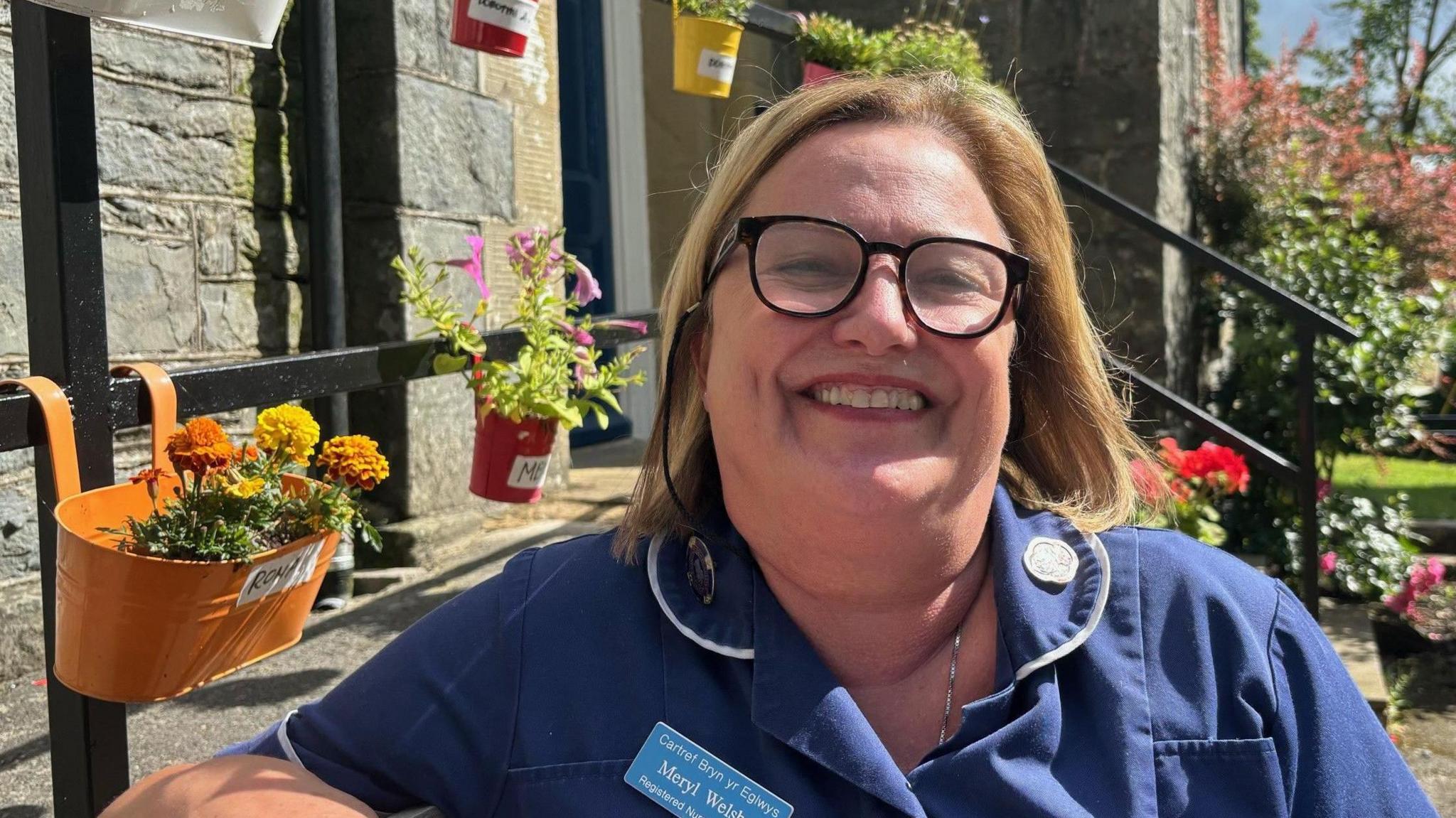 Meryl Welsby smiling and wearing a blue nurse's top sitting on a bench beside flowers in front of the home