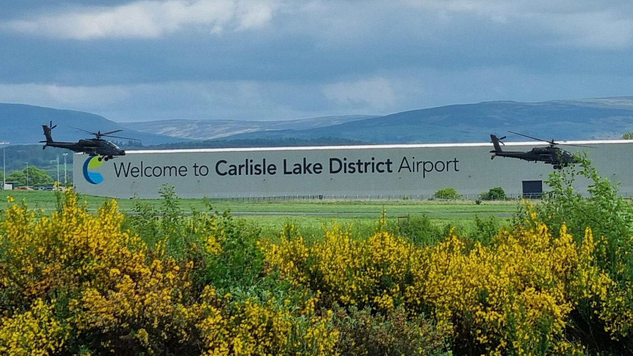 The words 'Carlisle Lake District Airport' are displayed on the side of a large single-storey white building. Mountains are seen in the background. Helicopters fly in front of the building