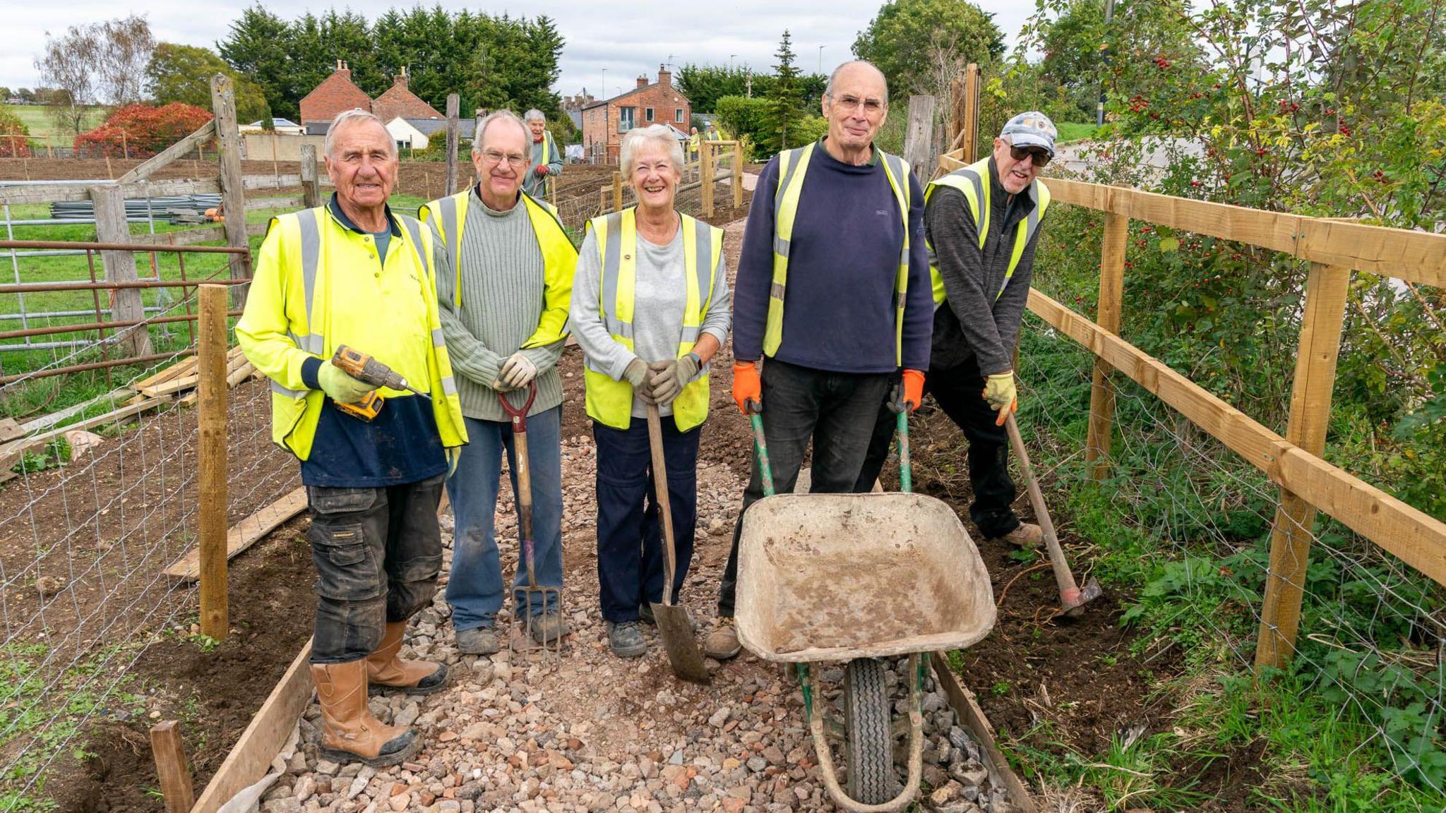 A group of five older people - four men and one woman - don yellow hi-vis jackets and construction boots as they do some community landscaping. They are standing on a new gravel path path near a wooden fence in the countryside.