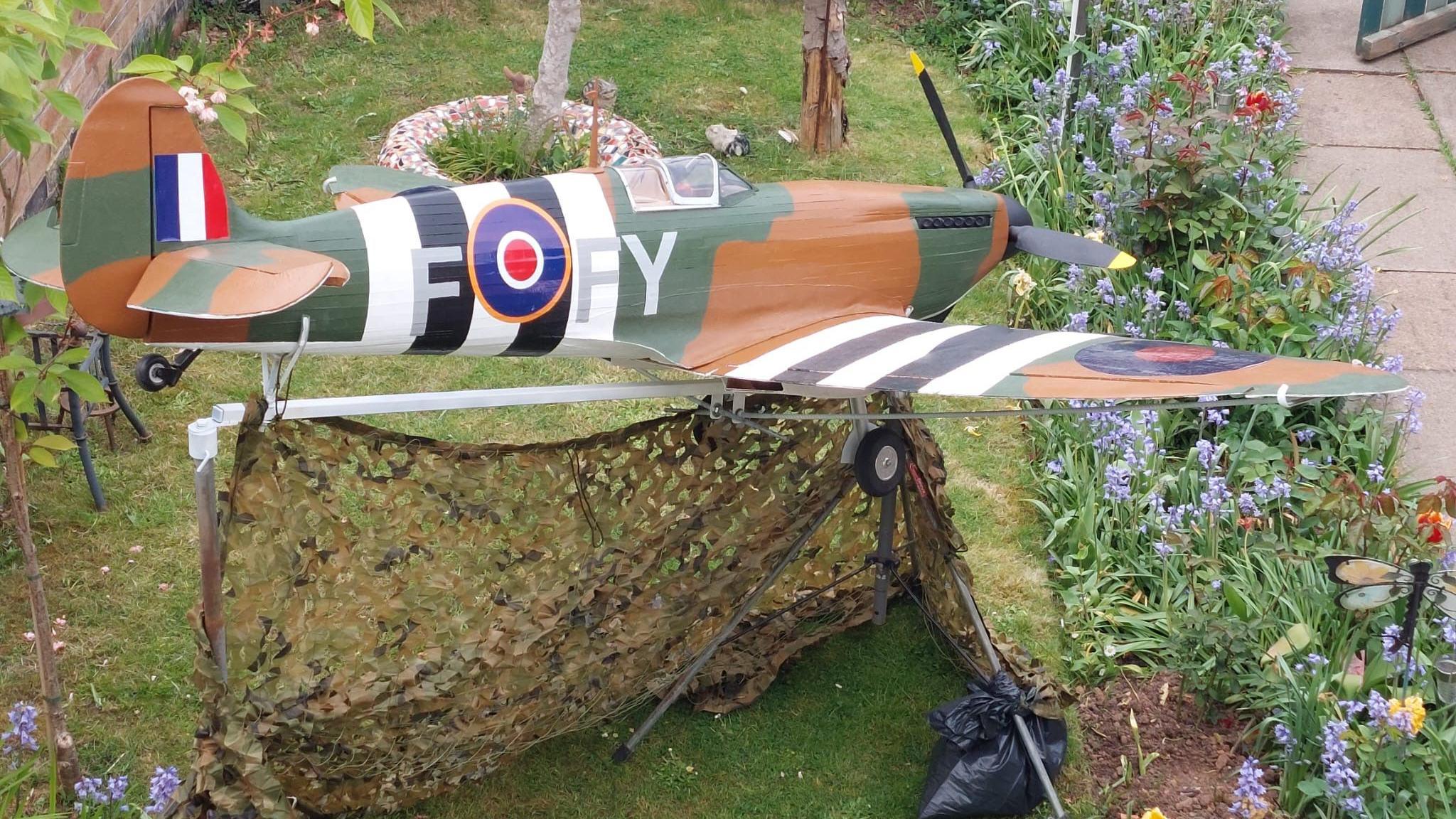 A large model Spitfire plane in a garden. The plane is dark green, white, black and brown with black propellers on its nose. It has an RAF circular logo on the side and camouflage netting underneath it. The picture is taken from above and behind it. There is a French flag on its tail, small wheels underneath it and a cockpit