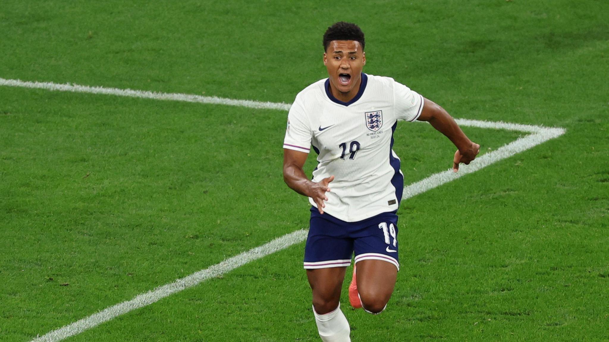 Ollie Watkins wearing the England White jersey and blue shorts running in celebration after scoring a goal