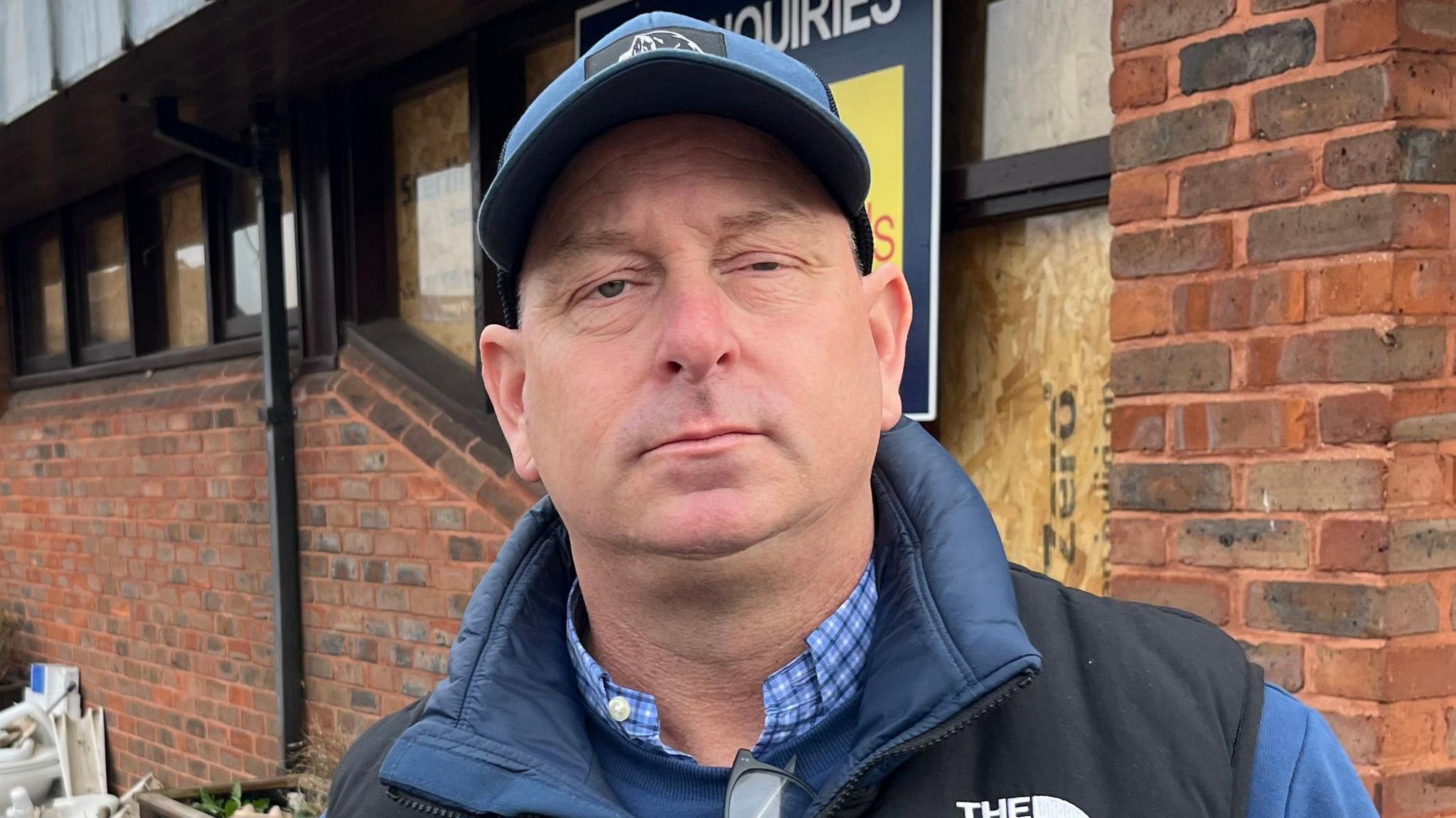 A man wearing a blue baseball cap, a blue body warmer over a blue top and a blue and white chequered shirt underneath. He is standing in front of a corner of an old brick pub with a large pile of debris and rubble to the left.
