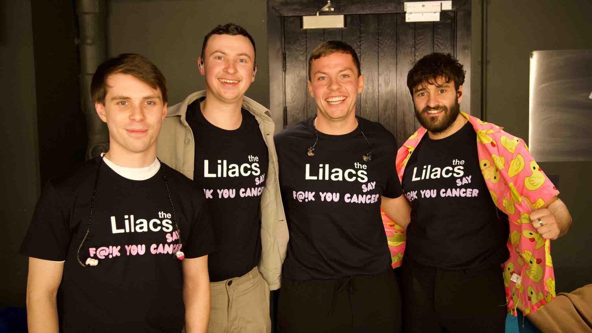 Four men in their 20s. The Lilacs pictured backstage before a show. All four bandmembers are wearing a t-shirt reading, 'the Lilacs, say f@1K you cancer'.