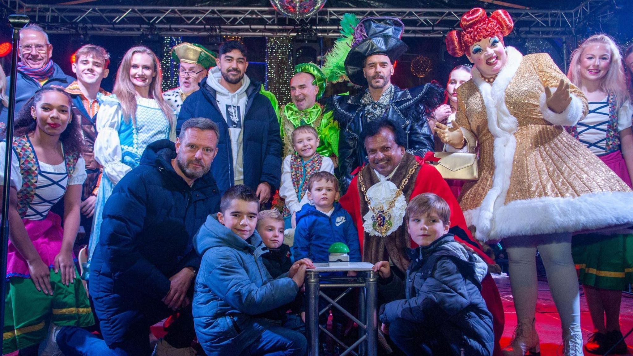 A large group of people stand on a stage in the centre of Ipswich. Among them is Ipswich Town FC chief executive Mark Ashton, who is kneeling on the front row, and panto star Louie Spence, who is at the back dressed in a vivid green outfit for his part in Jack and the Beanstalk at the Wolsey Theatre. There are other pantomime characters on the stage along with local childen