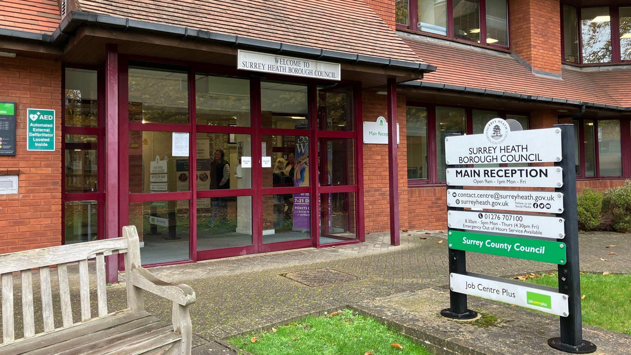 Entrance to Surrey Heath Borough Council in Camberley. It is a red-brick building with a wooden bench outside and a path and a grass verge.