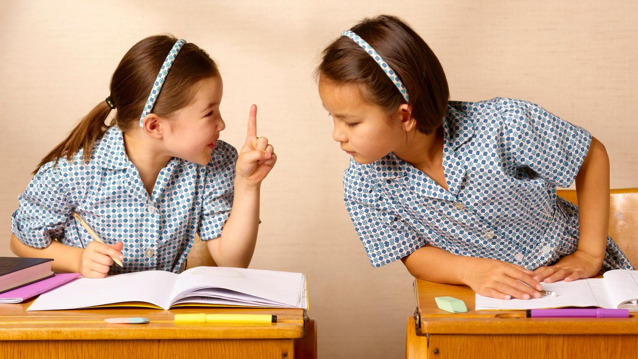 two children in blue spotty uniforms sitting at wooden desks, one trying to copy the work of the other, with the other telling her off for it