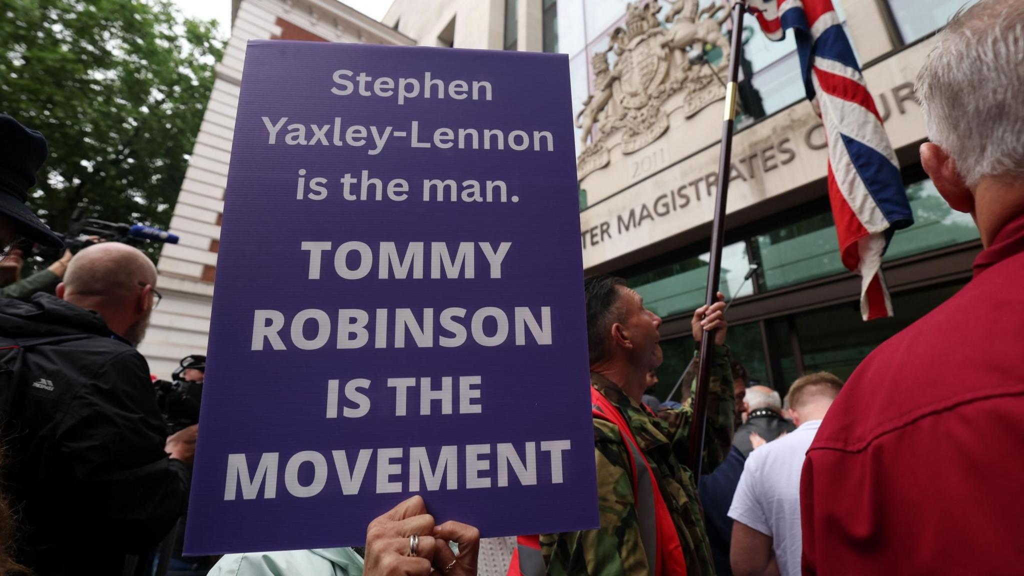 A purple placard with white writing on it saying "Stephen Yaxley-Lennon is the man. TOMMY ROBINSON IS THE MOVEMENT". A man holds a Union flag on a short pole. The entrance to Westminster Magistrates Court is visible behind them.