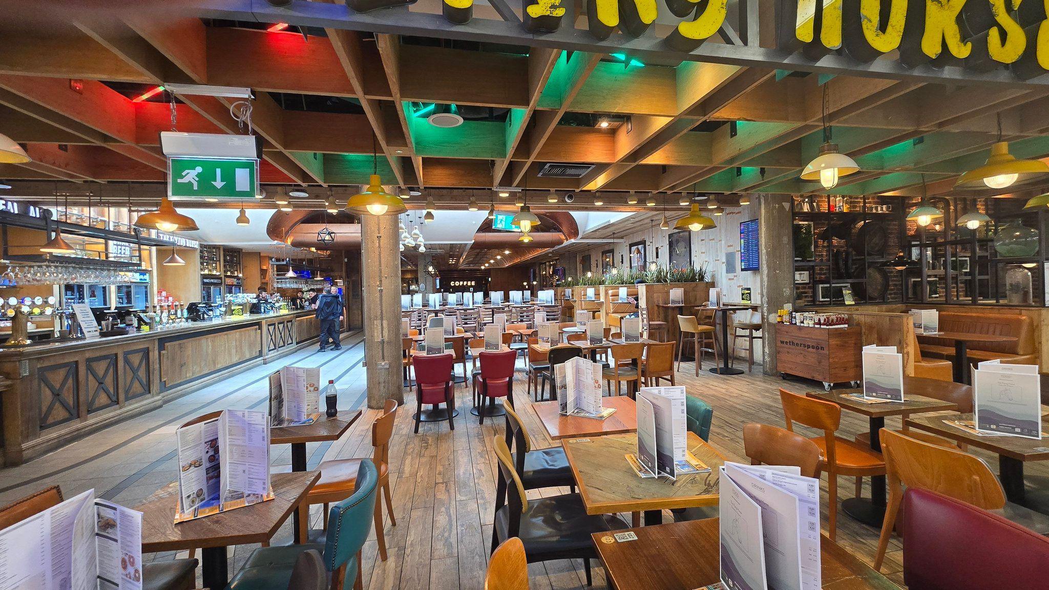 Interior of a large airport restaurant showing tables and chairs, menu displays, a bar counter and overhead lighting. Only a small number of people are visible near the bar area while much of the seating is empty.
