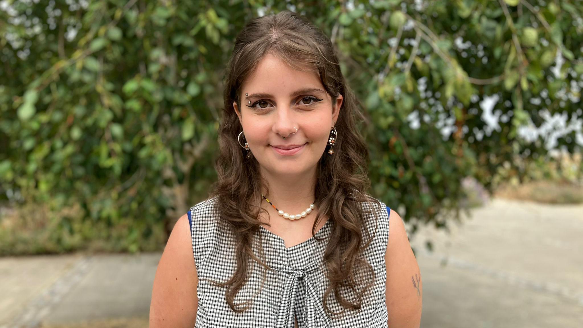 A student with long curly brown hair, a pearl necklace and a black and white squared top is standing in front of a tree in a park looking straight at the camera