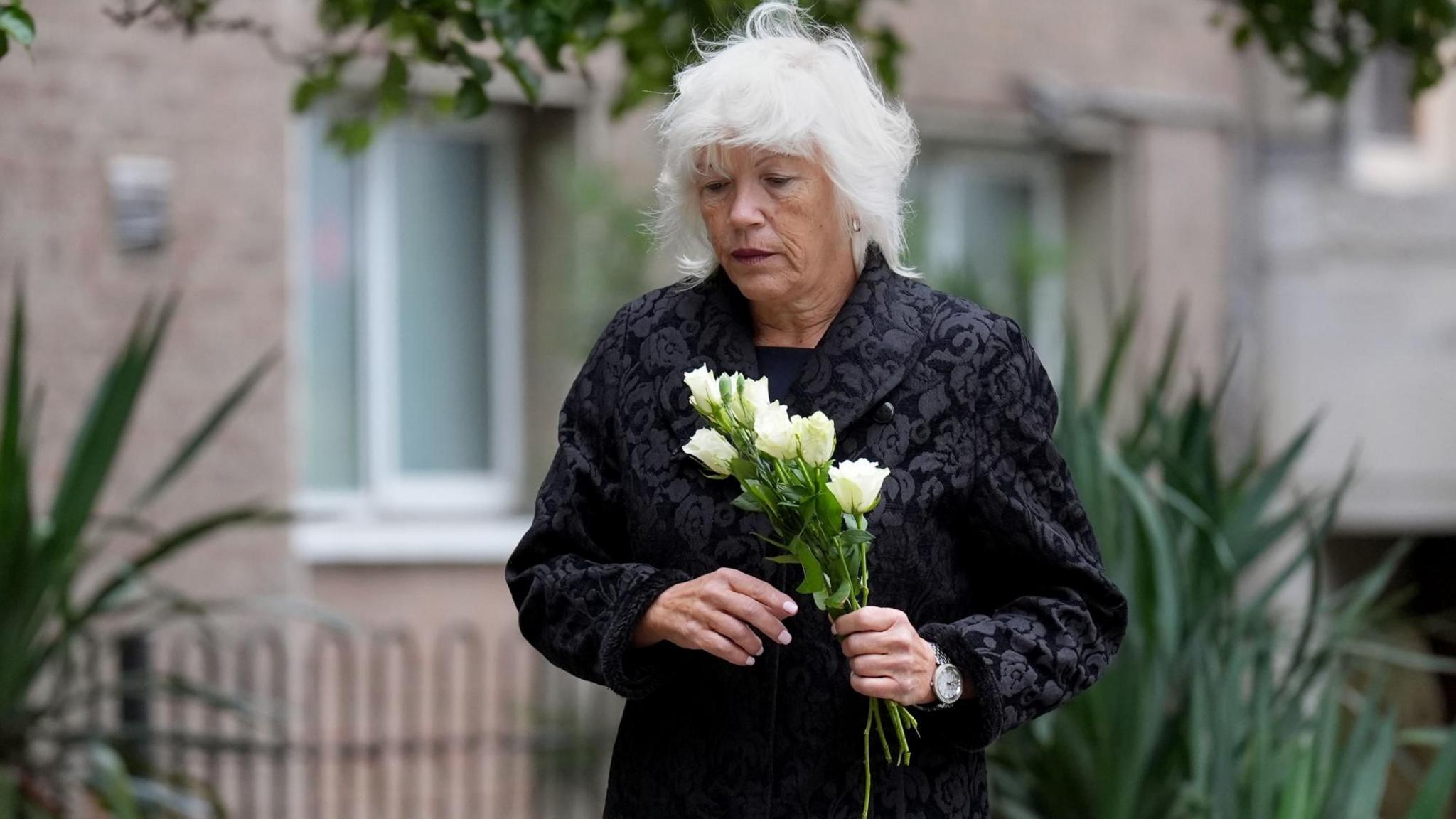 Melanie Leahy carrying white roses in her hand outside Chelmsford Civic Centre where the Lampard inquiry is being held. She is looking down and dressed in a black, long sleeved coat. Her hair is grey and shoulder length. She is about to place the flowers on a poster of her son placed on the ground alongisde photos of other people that have died under the care of mental health services in Essex