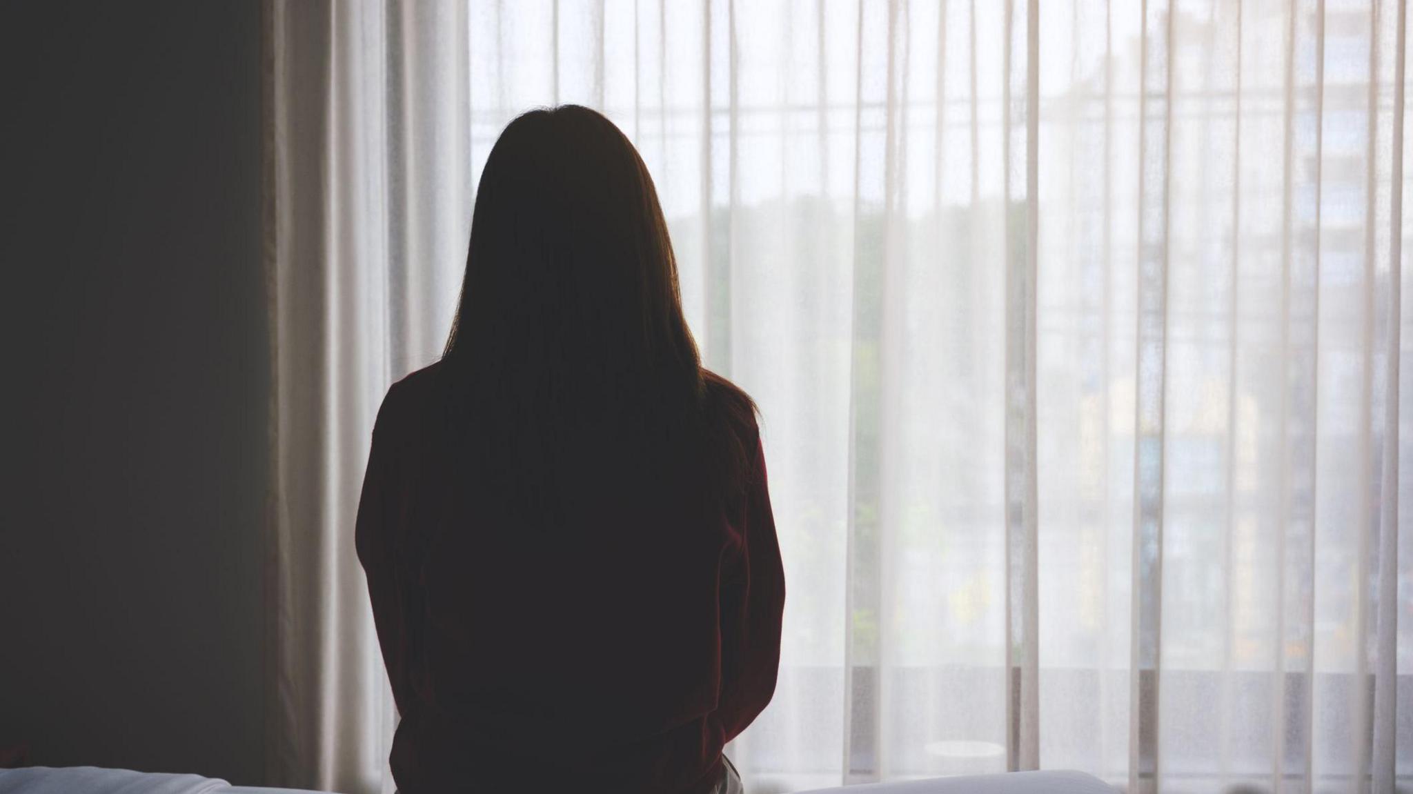 A woman, who is silhouetted, sits on a bed staring out of a window