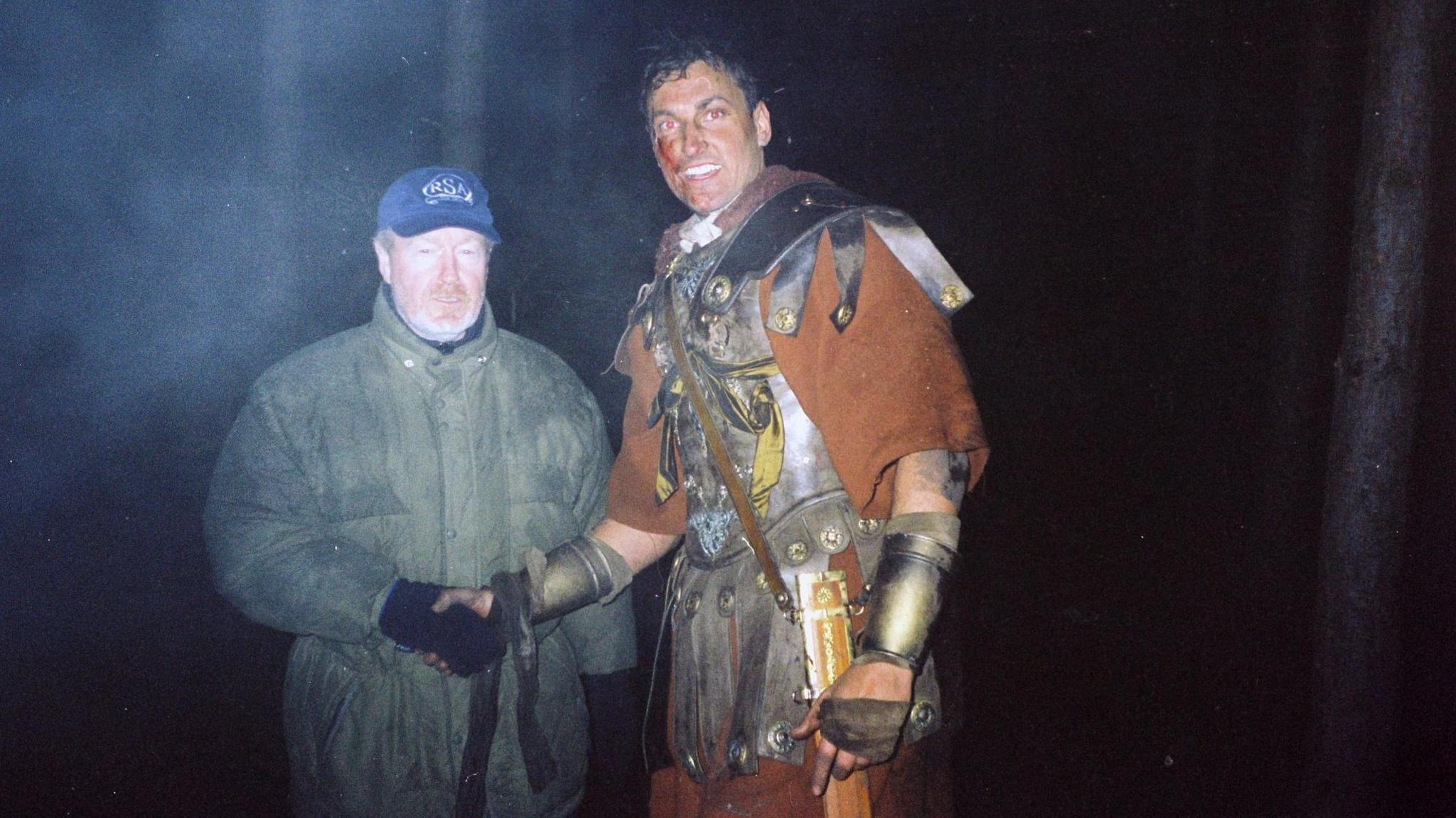 Andy Bowden, dressed as a Roman centurion shakes hands with director Ridley Scott. They are in woodland at night, with mist behind them.