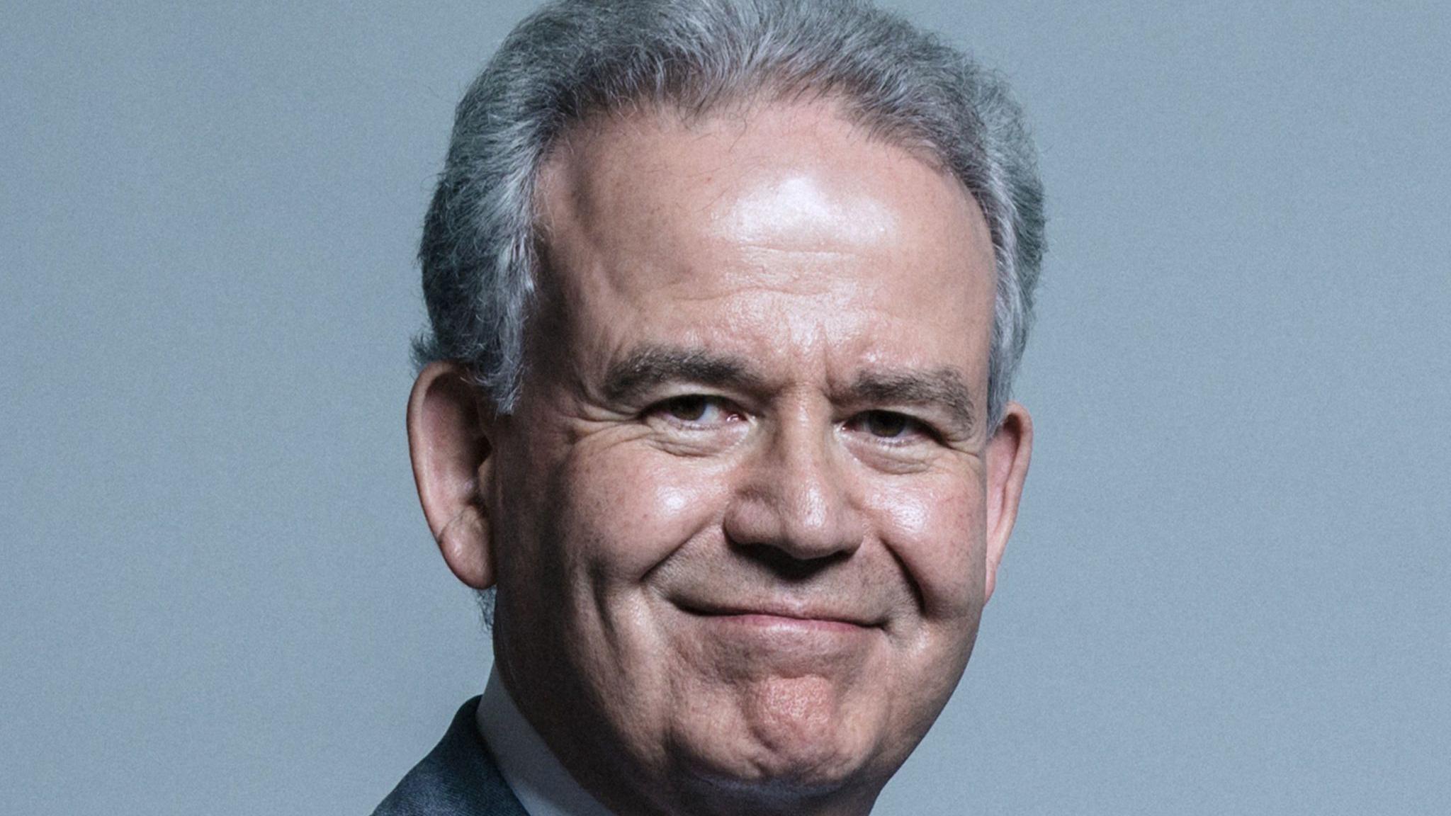 Sir Julian Lewis smiles at the camera against a light grey background. The MP wears a suit and has combed-back grey hair