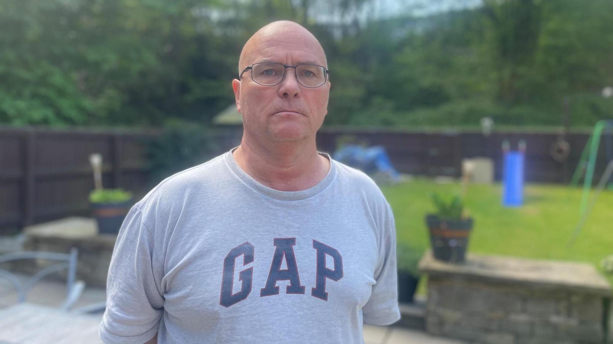 A cross stocky middle aged man with shaved hair wearing a grey t-shirt looks towards the camera