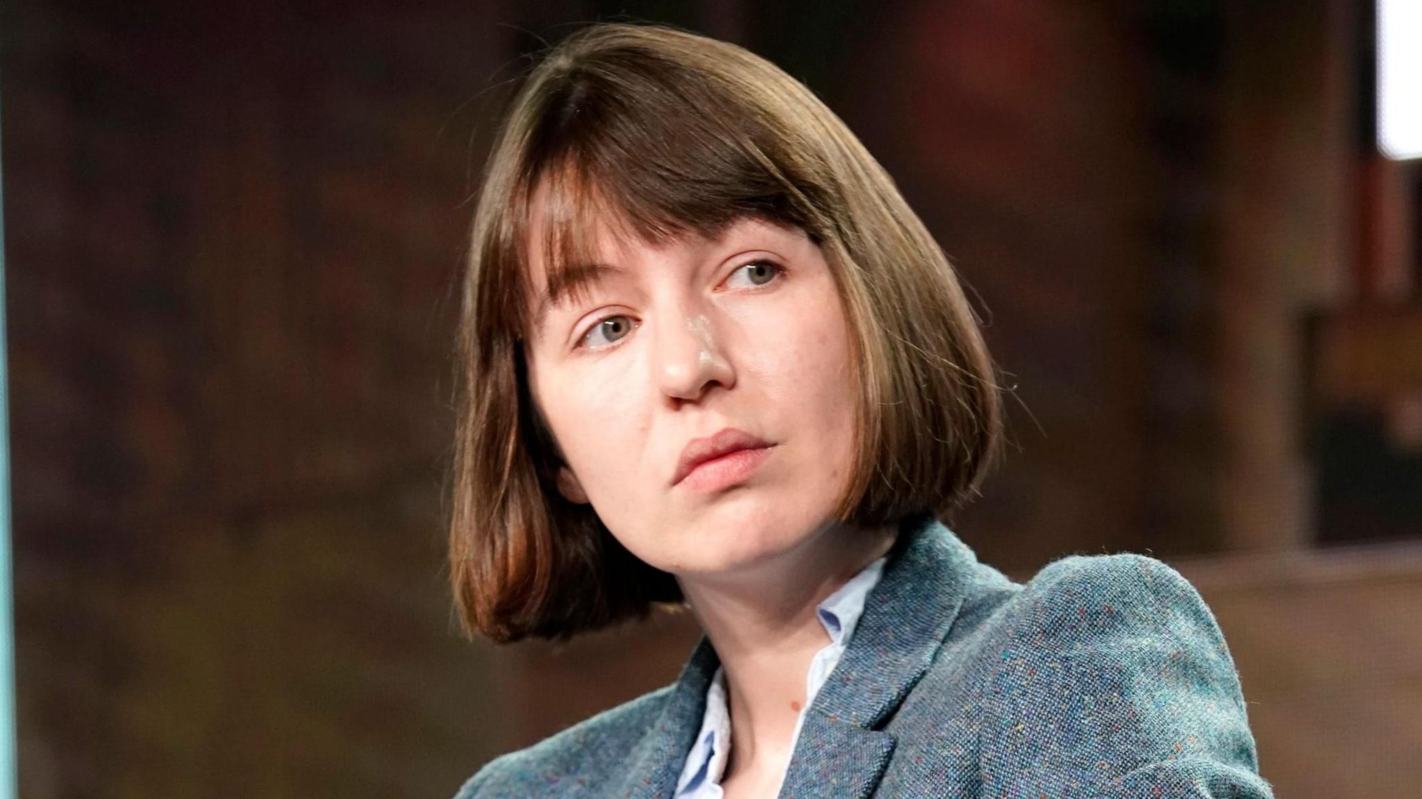 Author Sally Rooney looks sideways, wearing a blue jacket. She has a brown bob with a fringe