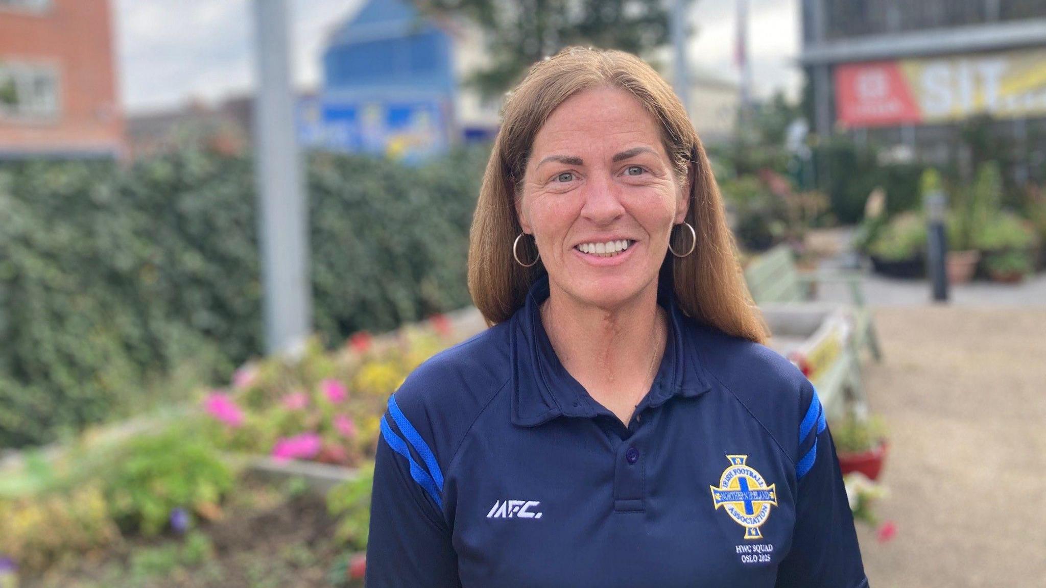 Louise McGonnell smiles at the camera. She has long brown hair, and is wearing hoop earrings. She is wearing a blue top with an Irish Football Association crest on it. Behind her are potted plants, a hedge, and a green bench.