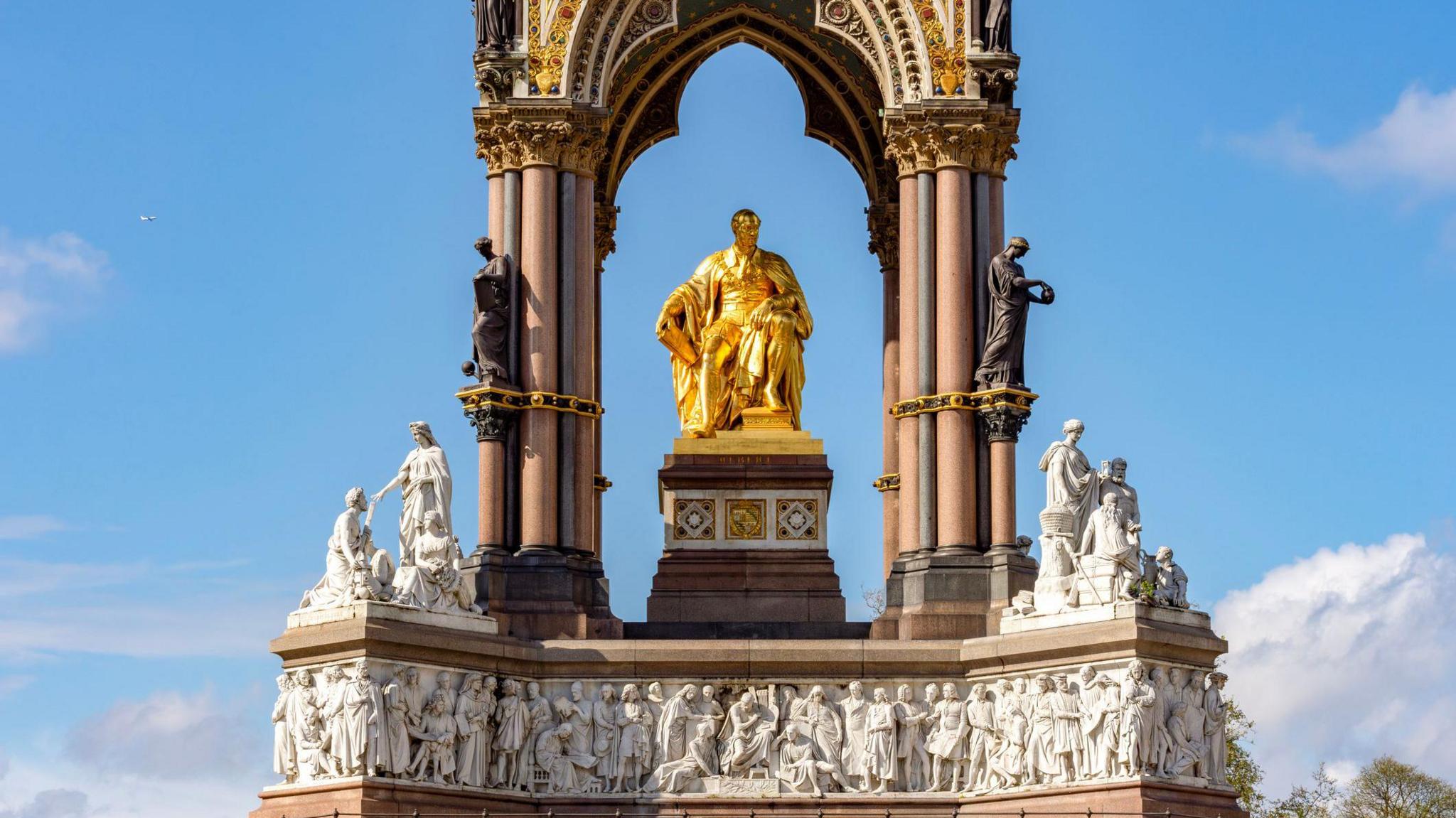 the Albert Memorial in Kensington Gardens, London. At its center is a gilded statue of Prince Albert seated under a Gothic canopy. The memorial features ornate columns and a base adorned with white marble statues. The clear blue sky in the backdrop.