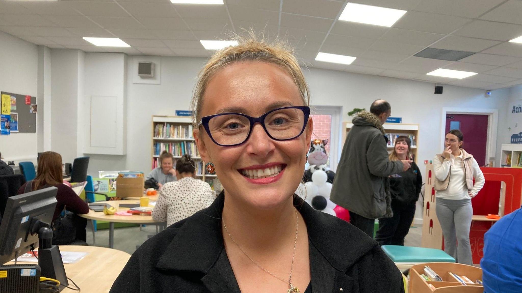 A close-up photo of a white-skinned lady with blond hair and wearing black-framed glasses in the library with people standing and sitting in the background. 