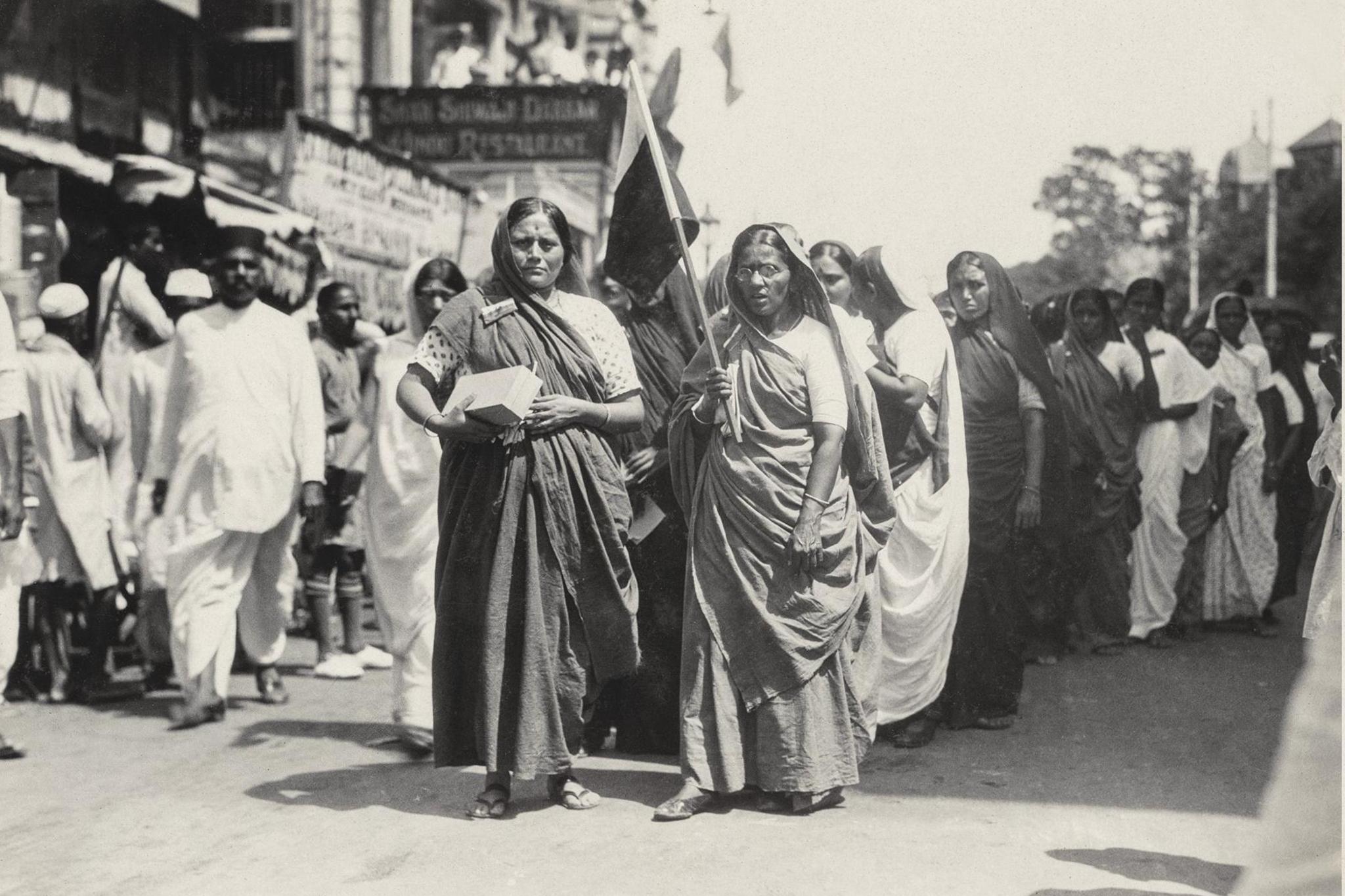 Two women in saris and with their heads covered, are seen leading a boycott procession in India. The photo, which is monochrome, shows them walking along a busy street. The woman on the right is wearing glasses and is holding a provisional national flag.