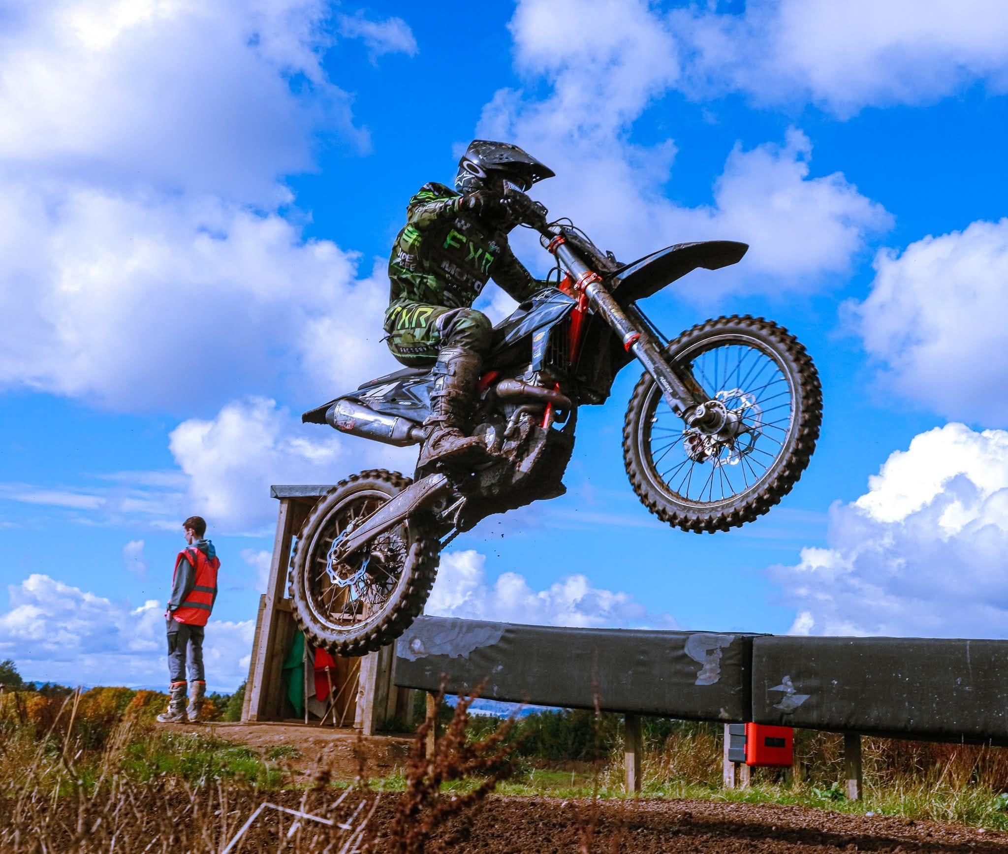 A dynamic moment as a person, riding a dirt bike, is caught mid-air during a jump. The rider is equipped with protective gear, including a helmet and clothing featuring green and black patterns. In the background, there's another individual wearing an orange vest, possibly an official, standing near a small structure.