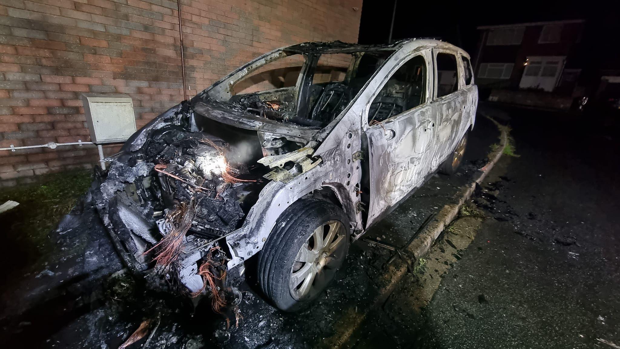 Burnt car, with windows blown out, engine charred and white paint turned grey and black. The car is near a wall.