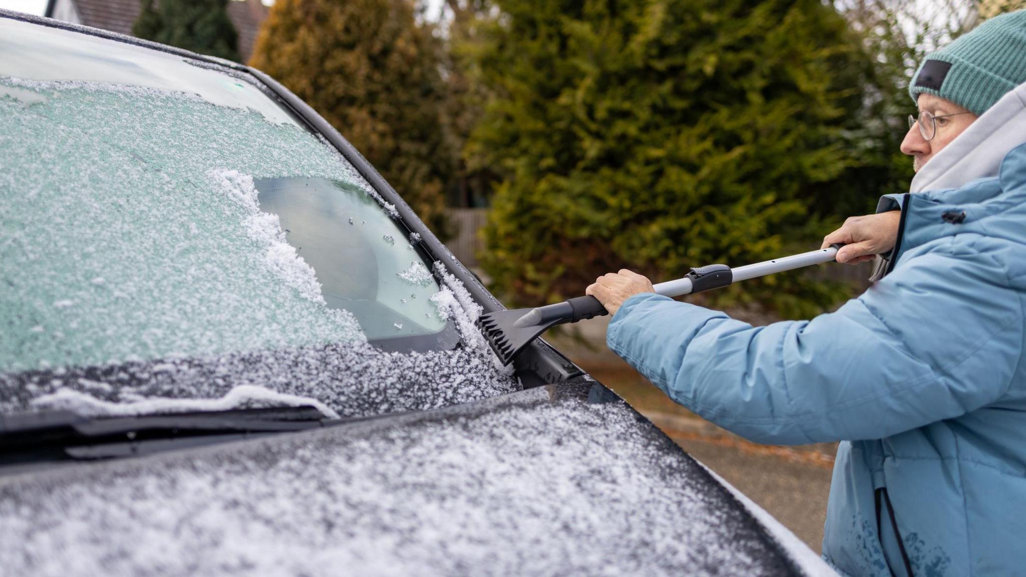 A man in a blue coat and a blue hat scraping ice of a car.