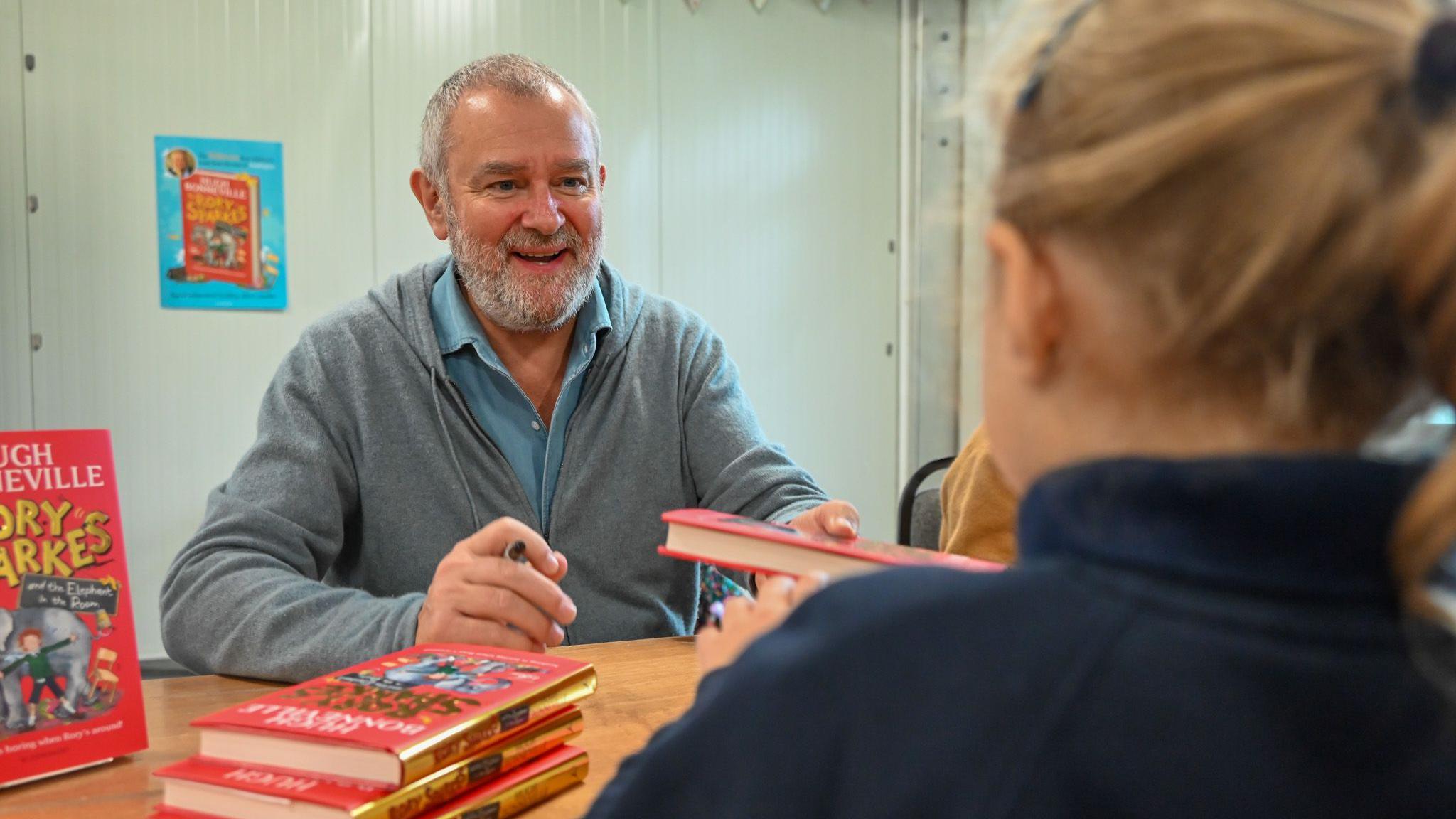 Hugh Bonneville smiles and talks as he hands a red bound hardback book back to a young girl with blonde hair, who has her back to the camera and is wearing a navy polo shirt. Bonneville is holding a black permanent marker in his other hand, and a small stack of his books lies on the wooden table in front of him.