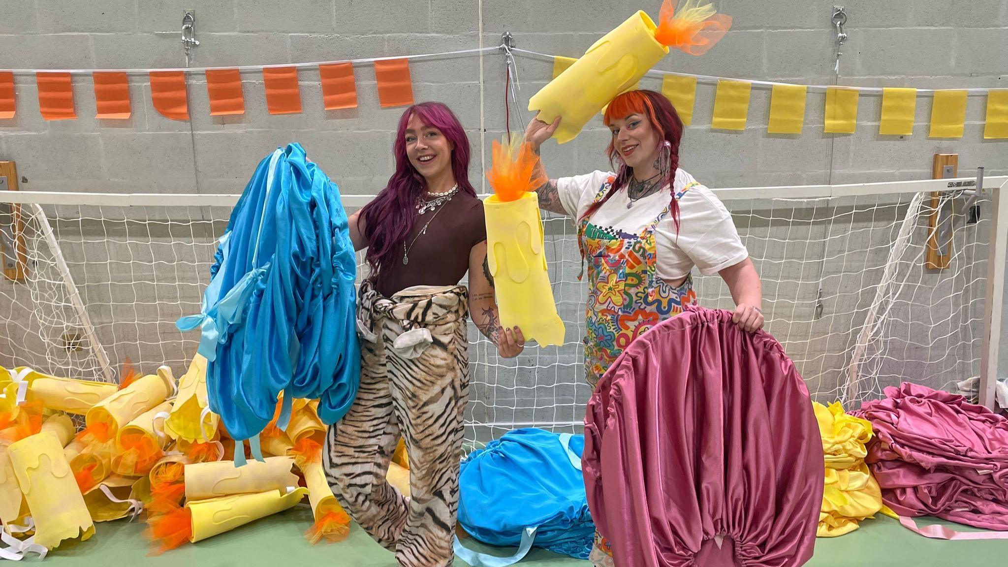 Two women standing in a hall holding up homemade bright yellow flaming candles and blue and pink costumes. The woman on the left is wearing animal print trousers and a brown top and has long dark pink-tinted hair. The woman on the right has red dyed long hair in pigtails and is wearing a white T-shirt and bright floral dungarees. Piles of candles and costumes are behind the pair, in a mouth of a football goal.