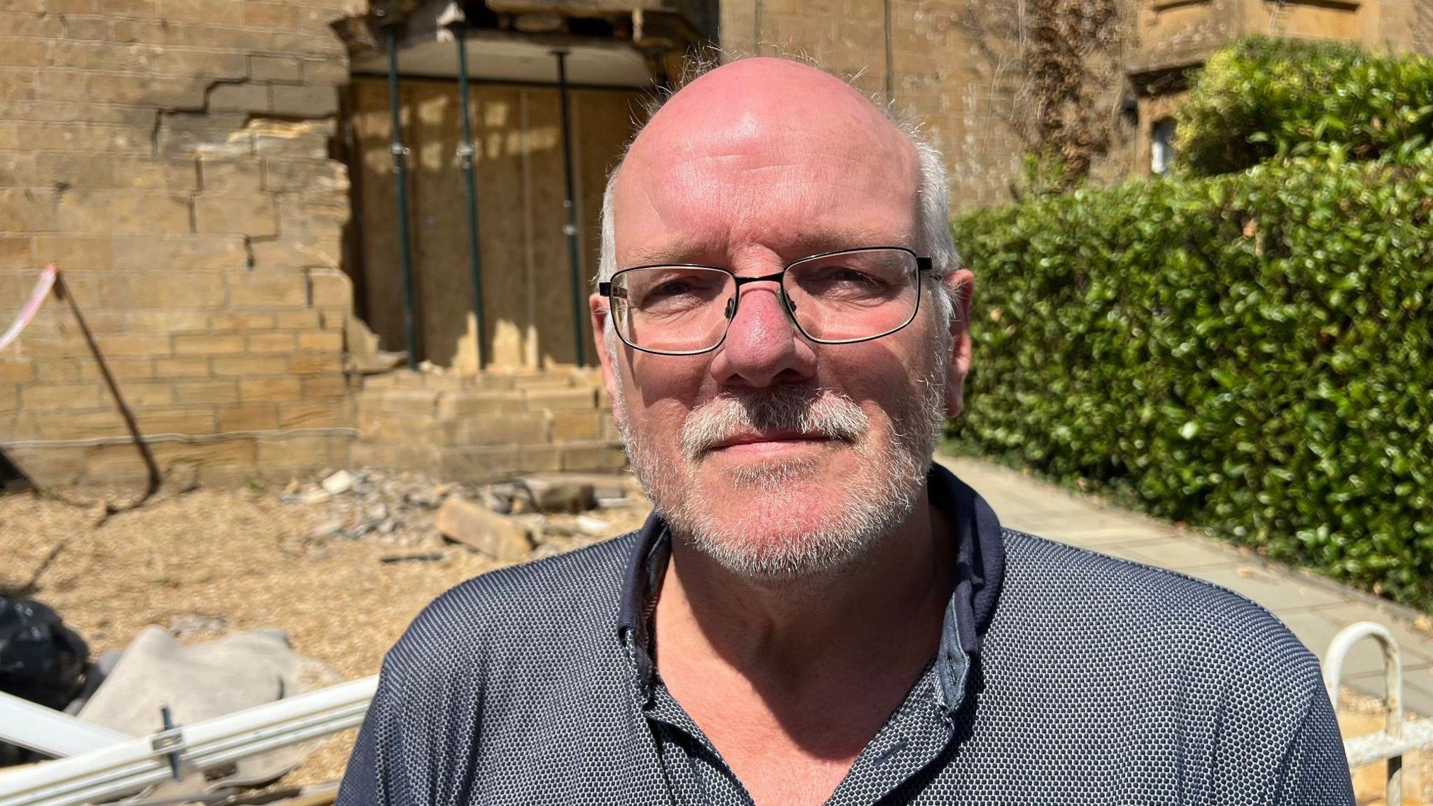 Marc looks at the camera with a small smiles. He has grey hair, and wears a grey shirt. Behind him is the hole in the living room window, with debris strewn across the garden.