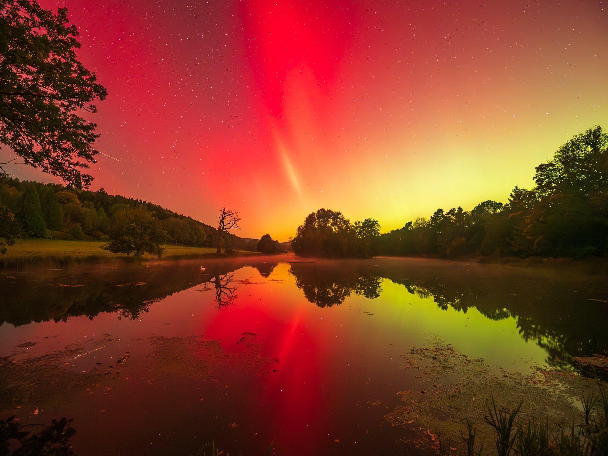 Northern Lights over Walcot Lake in Shropshire. Red and yellow colours fill the sky, reflecting in the lake below, which is fringed with a wood and fields