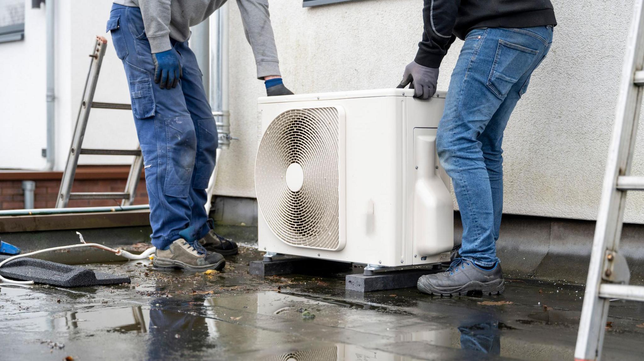 Two technicians installing an outdoor heat pump unit on a wet surface. They are wearing jeans and working boots and are holding the white box with a circular vent on the side. You can only see their torso and legs in the image.