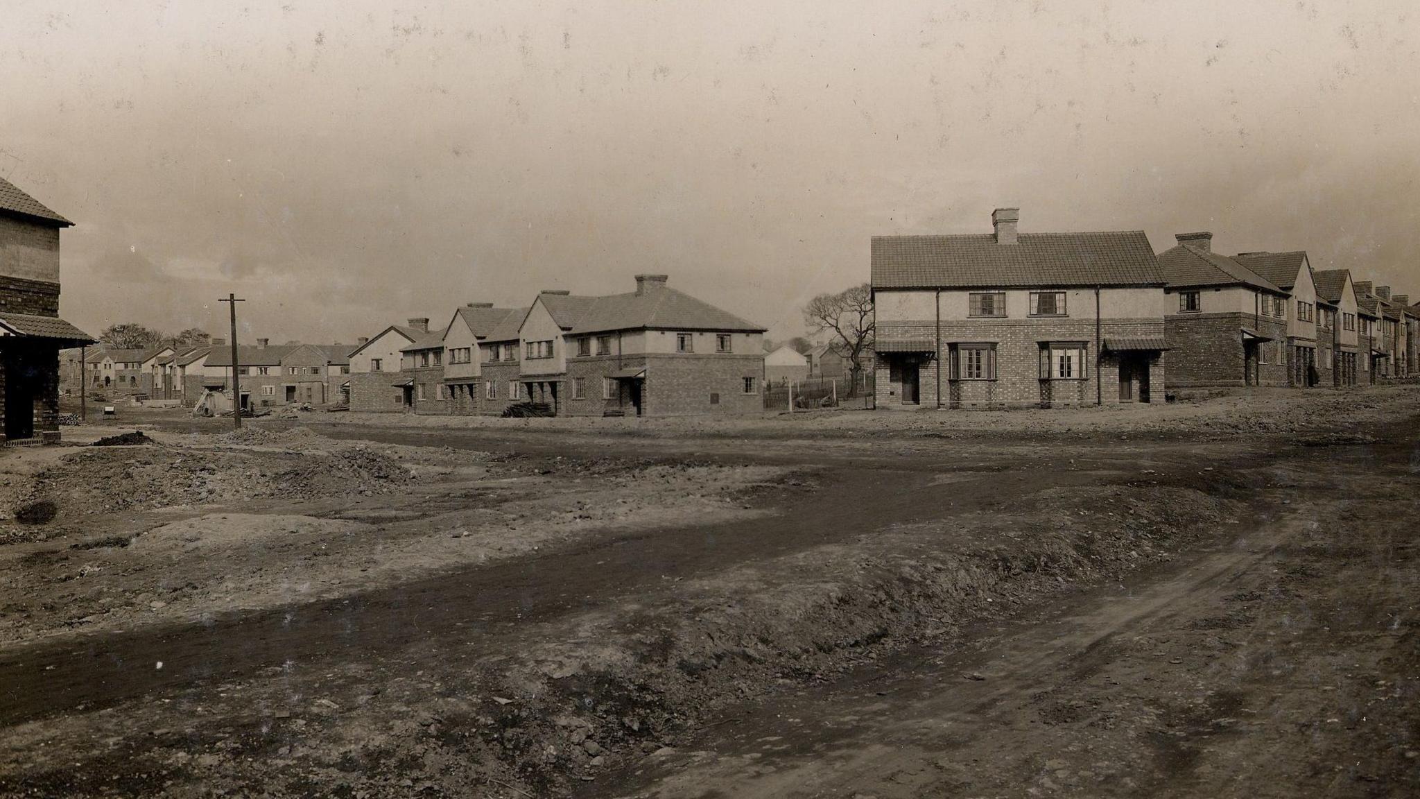 A black and white picture showing houses which are two-storey \and are mainly semi-detached. In the front of the photo is a large area of ground which is empty. 