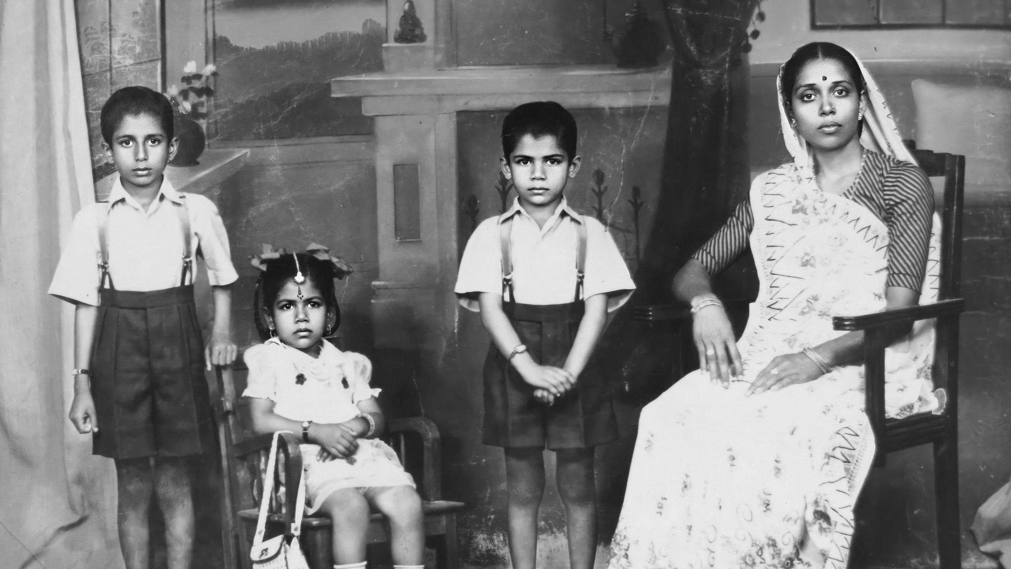 A black and white family portrait with four of Tara Mistry's family members. On the left is a young boy who is standing up wearing a short-sleeved shirt and black shorts. To his left, a young Tara Mistry is sitting down on a chair wearing a white dress. In the middle is another boy, standing and also wearing a shirt and shorts. On the far right is an older woman who is sitting down wearing a saree.